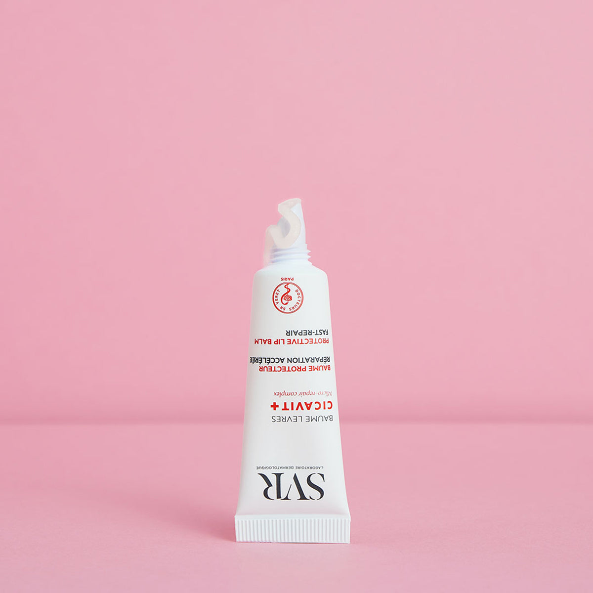 Tube de baume réparateur SVR Cicavit+ Lèvres blanc avec une pointe de crème sortant, sur fond rose pastel uni.
