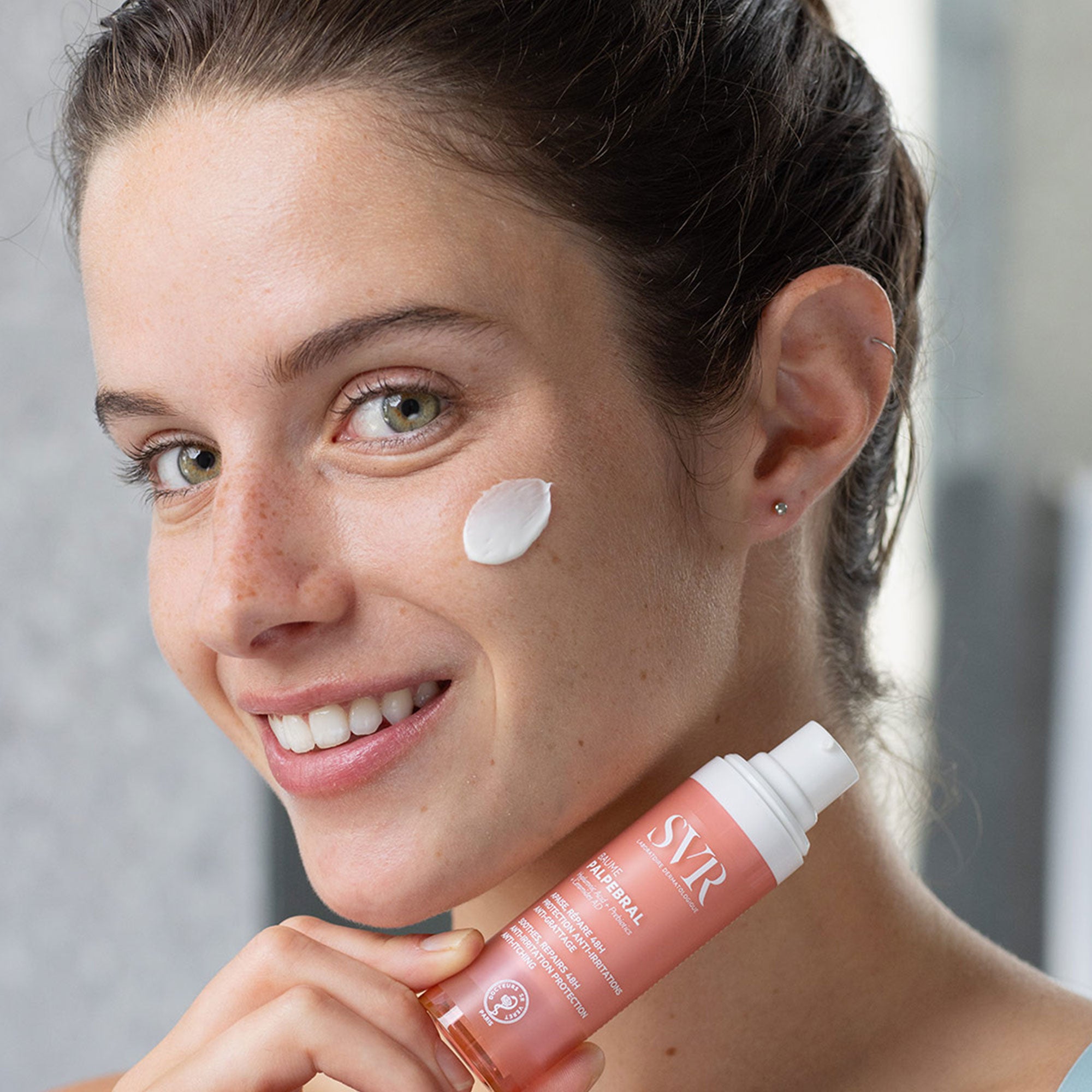 Femme souriante appliquant une noisette de baume blanc SVR Palpebral sur sa joue, flacon rose en main.