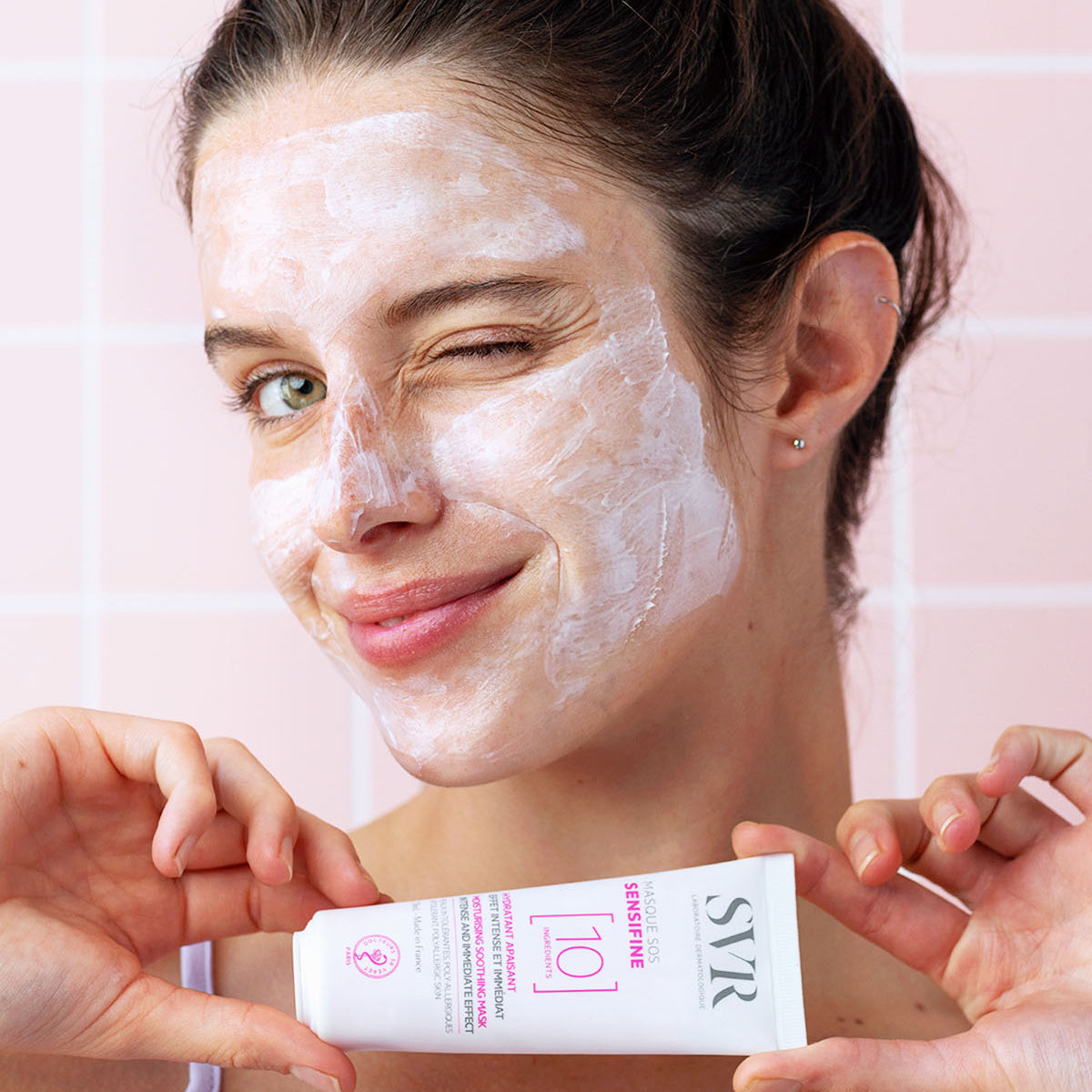 Femme souriante avec un masque visage blanc SVR Sensifine SOS et le tube de produit en main devant un carrelage rose.
