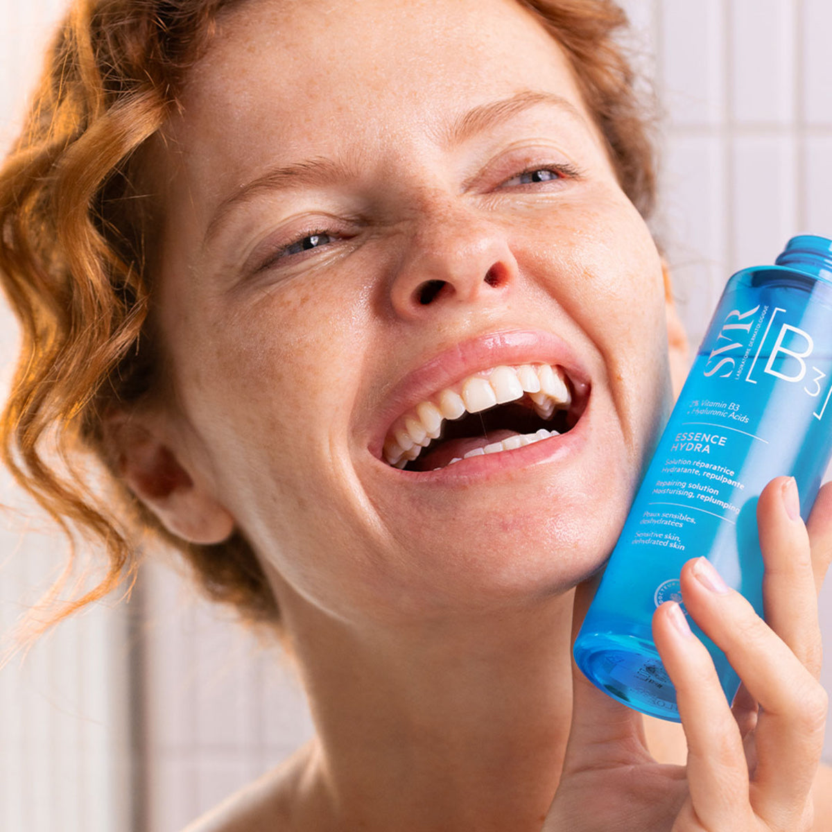Femme souriante tenant un flacon bleu SVR B3 Essence Hydra pour peaux sensibles et déshydratées.