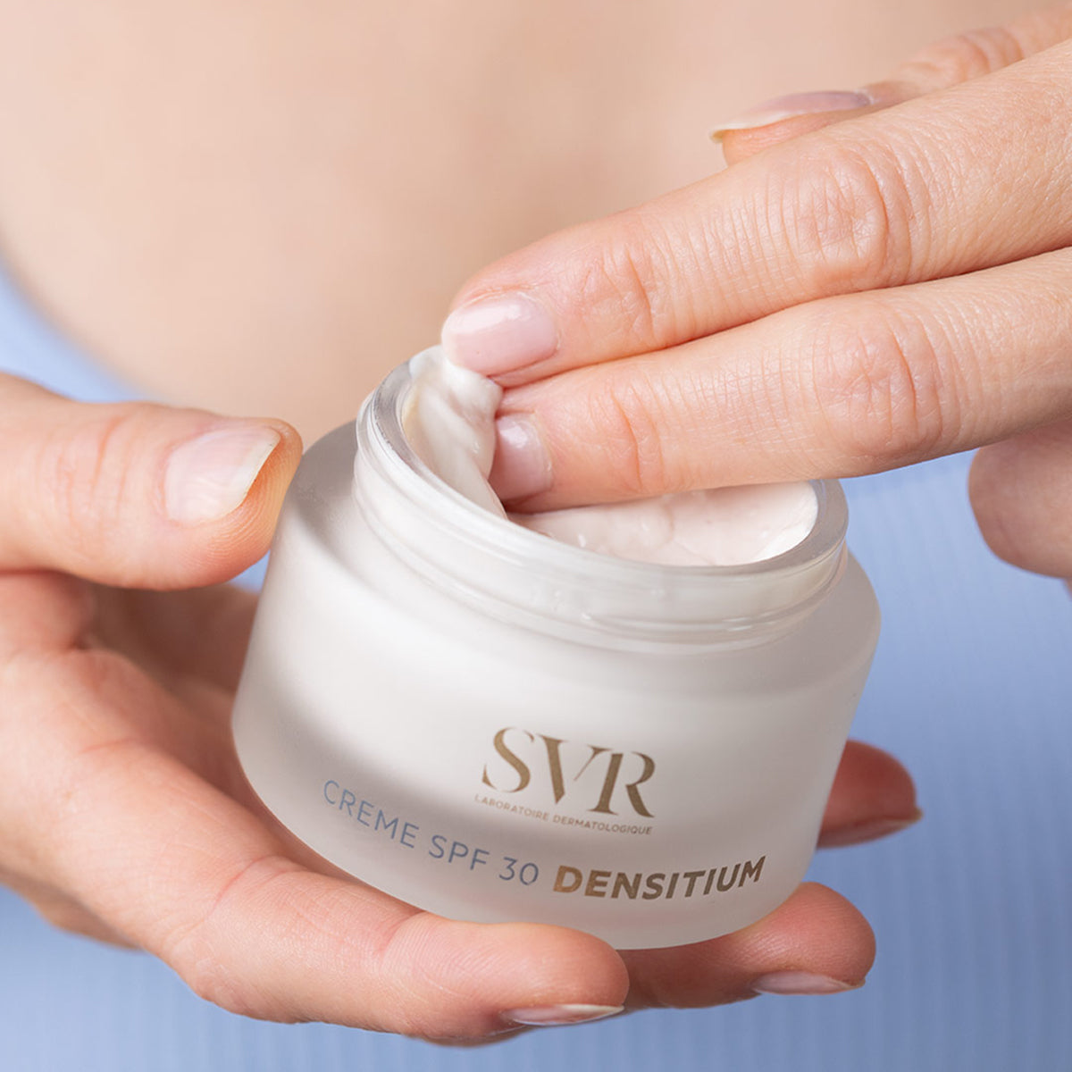 Hands holding a frosted glass jar of SVR DENSITIUM Crème SPF30 with a finger scooping the creamy white moisturizer.