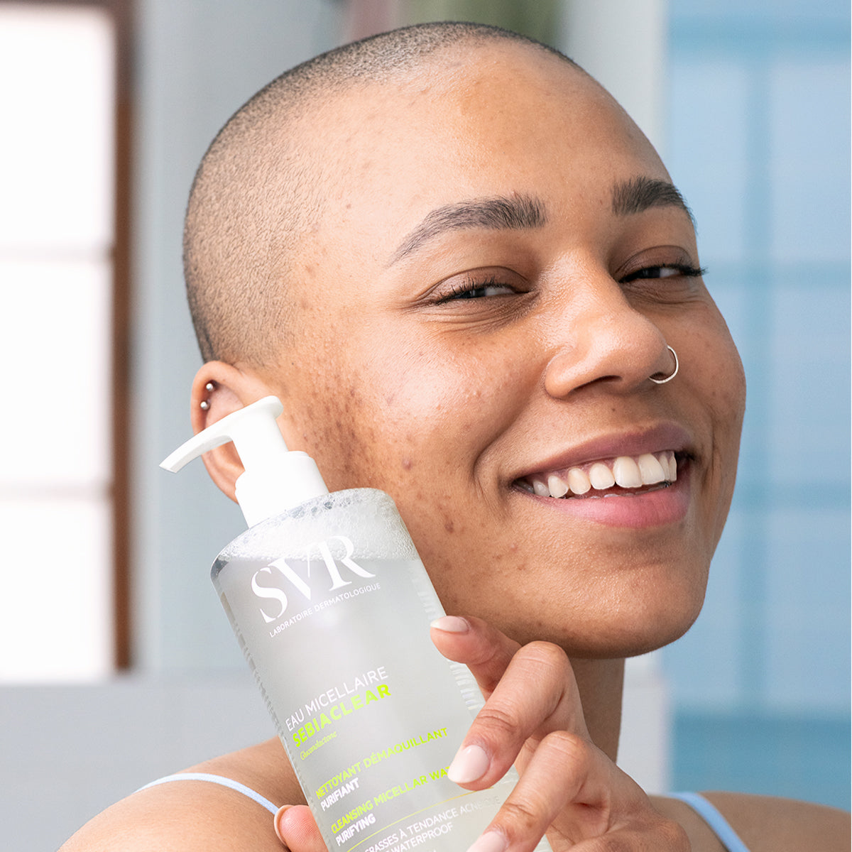 Femme souriante tenant un flacon pompe SVR Sebiaclear Eau Micellaire transparent pour peaux sensibles et acnéiques.