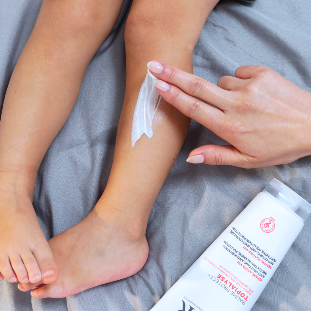 Main appliquant le baume réparateur SVR TOPIALYSE sur la jambe d'un enfant posée sur un drap gris.