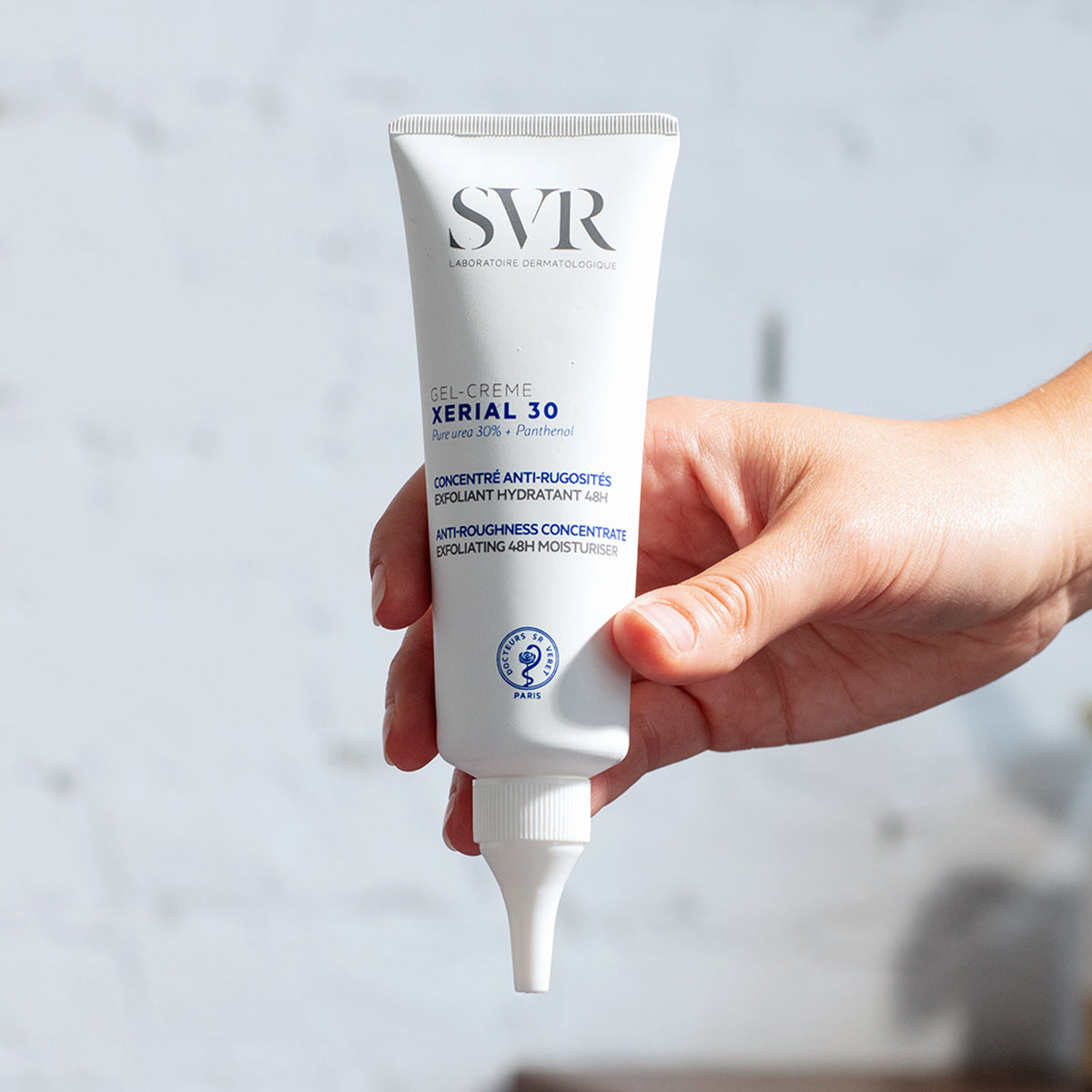 Tube blanc de gel-crème SVR Xerial 30 tenu en main, soin exfoliant hydratant pour peaux rugueuses avec embout applicateur précis.