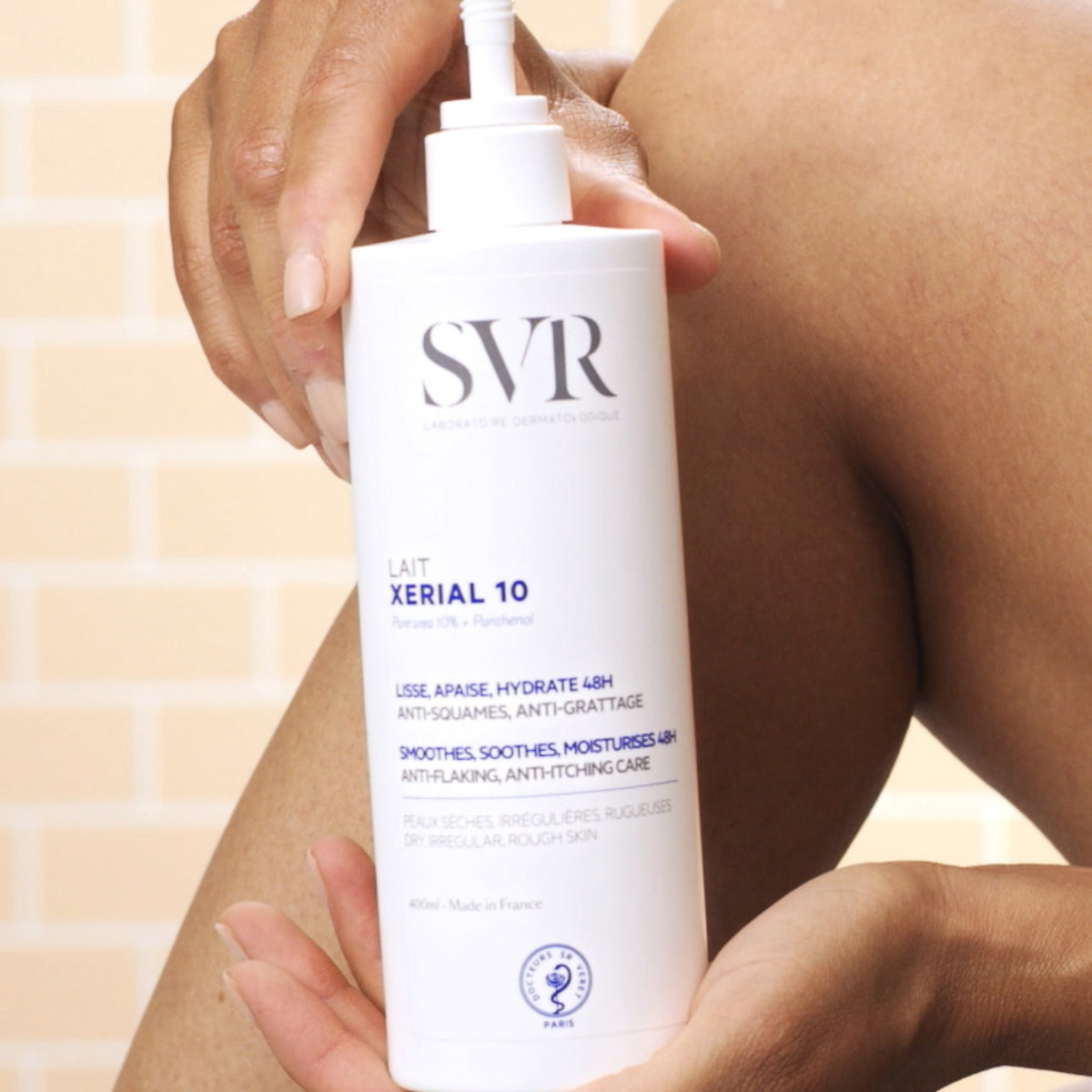 Flacon pompe blanc de lait hydratant SVR Xerial 10 de 400ml tenu à la main devant une jambe pour le soin des peaux sèches.