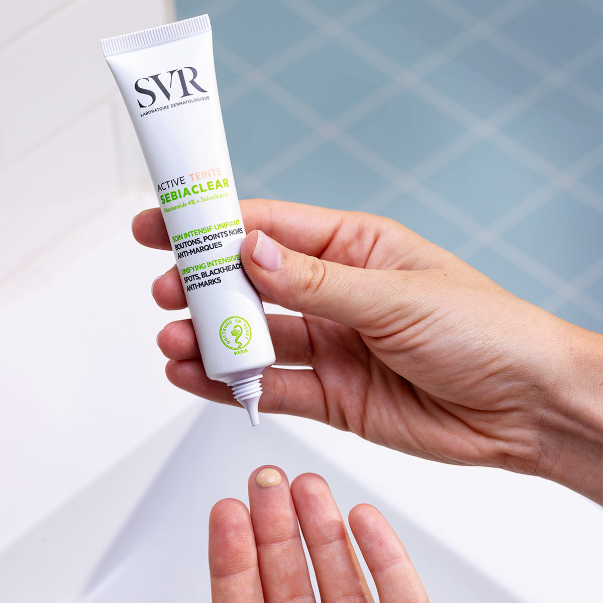 Hand holding a white tube of SVR Sebiaclear Active Teinté, dispensing a drop of tinted facial cream onto a fingertip.