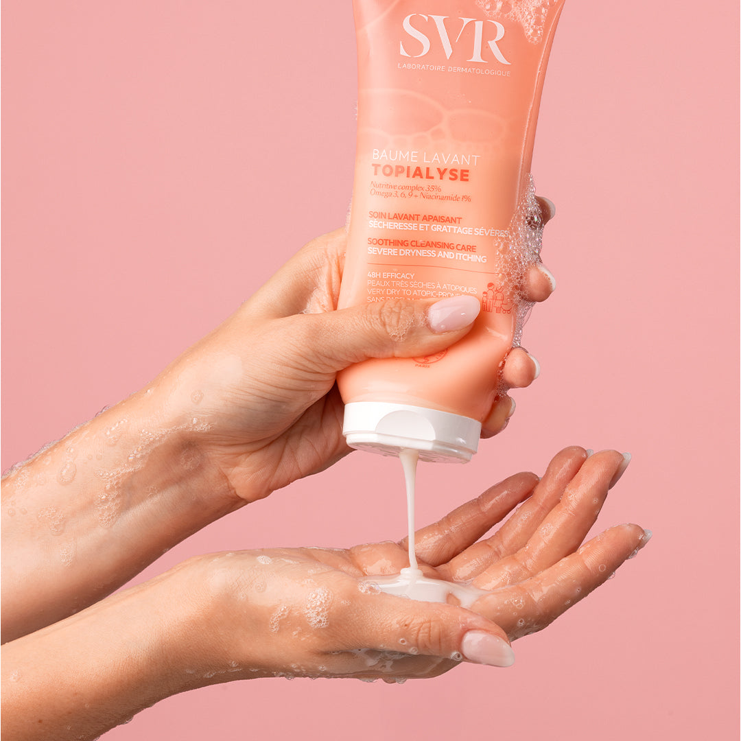 Tube de baume lavant SVR Topialyse rose pressé au-dessus d'une main, libérant une texture crémeuse sur fond rose.