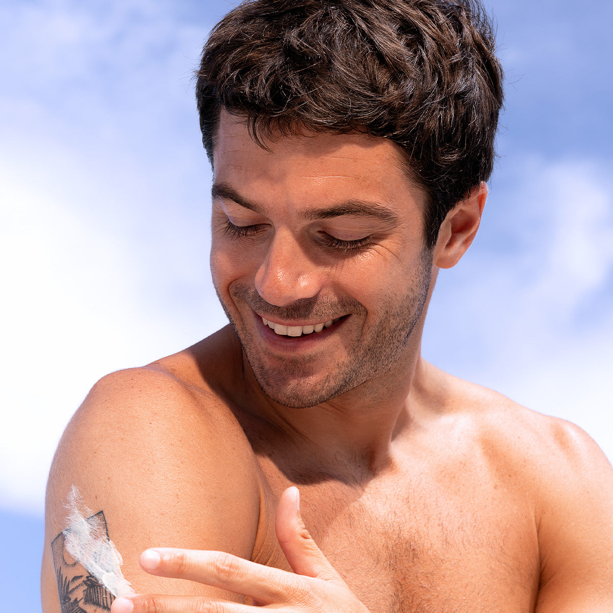 Homme souriant appliquant la crème réparatrice Cicavit+ SPF50+ sur son épaule tatouée sous un ciel bleu ensoleillé.