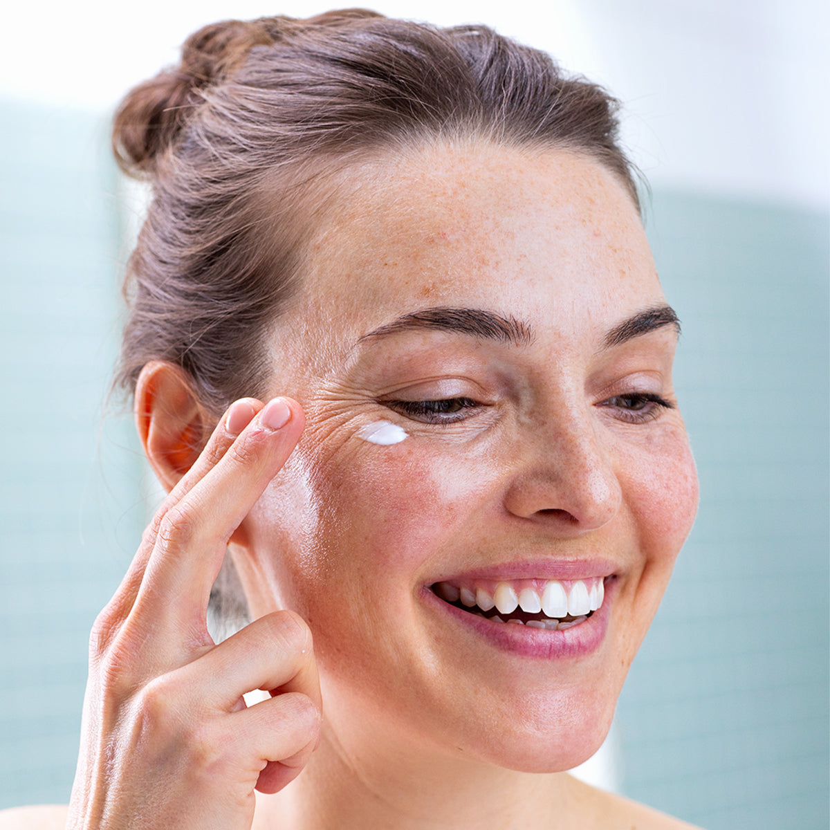 Femme souriante appliquant une noisette de crème blanche onctueuse sur le contour de ses yeux sensibles et irrités.