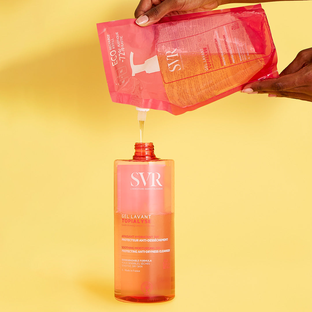 Hands pouring SVR TOPIALYSE Gel Lavant refill pouch into a clear 1L pump bottle against a yellow background.