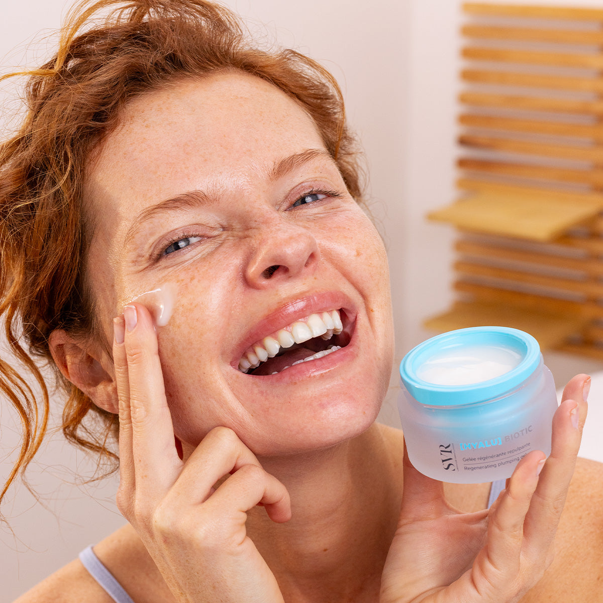 Smiling woman applying SVR Hyalu Biotic regenerating plumping gel from a blue and frosted glass jar.