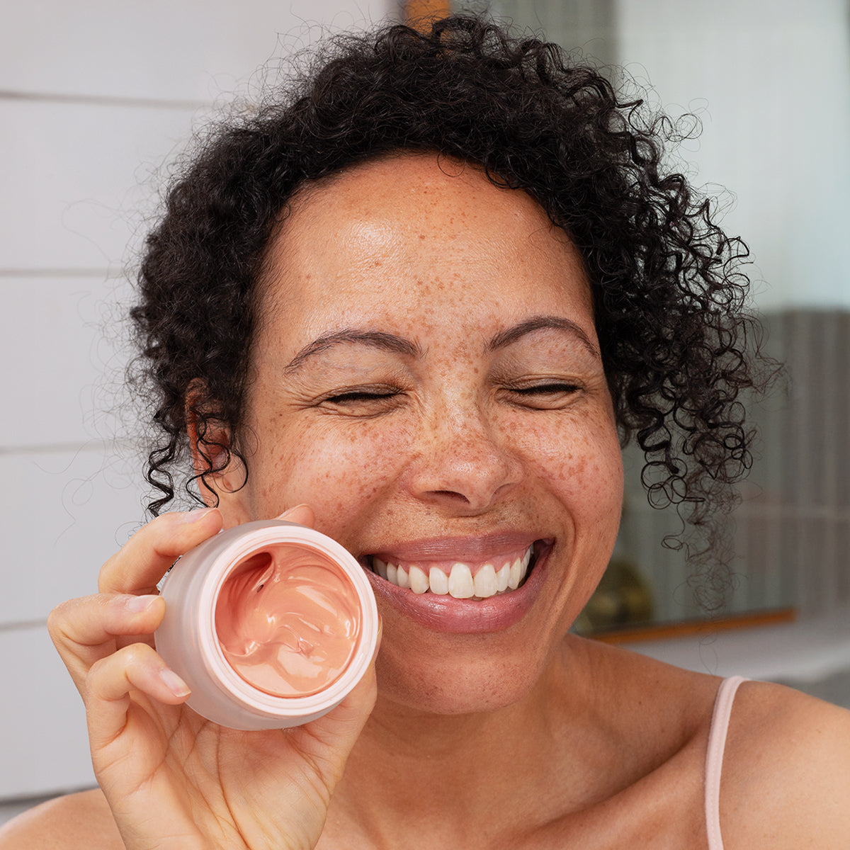 Smiling woman holding a jar of Densitium Rose Éclat cream with a soft pink, creamy texture.