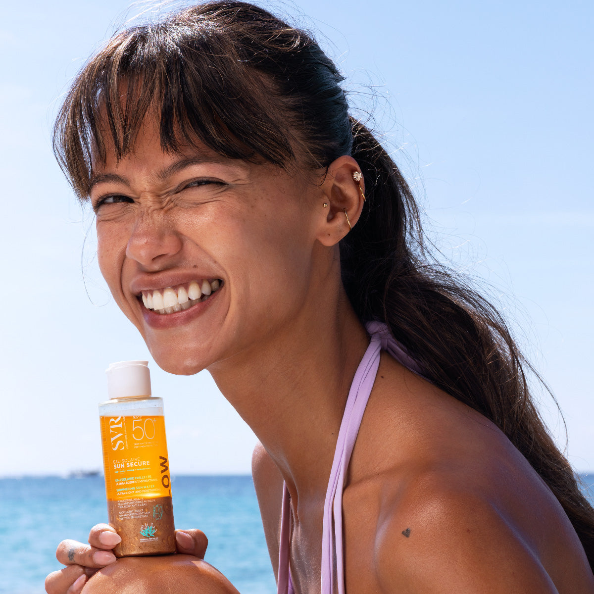 Smiling woman holding a bottle of SVR SUN SECURE Eau Solaire GLOW SPF50+ sunscreen against a sunny beach background.