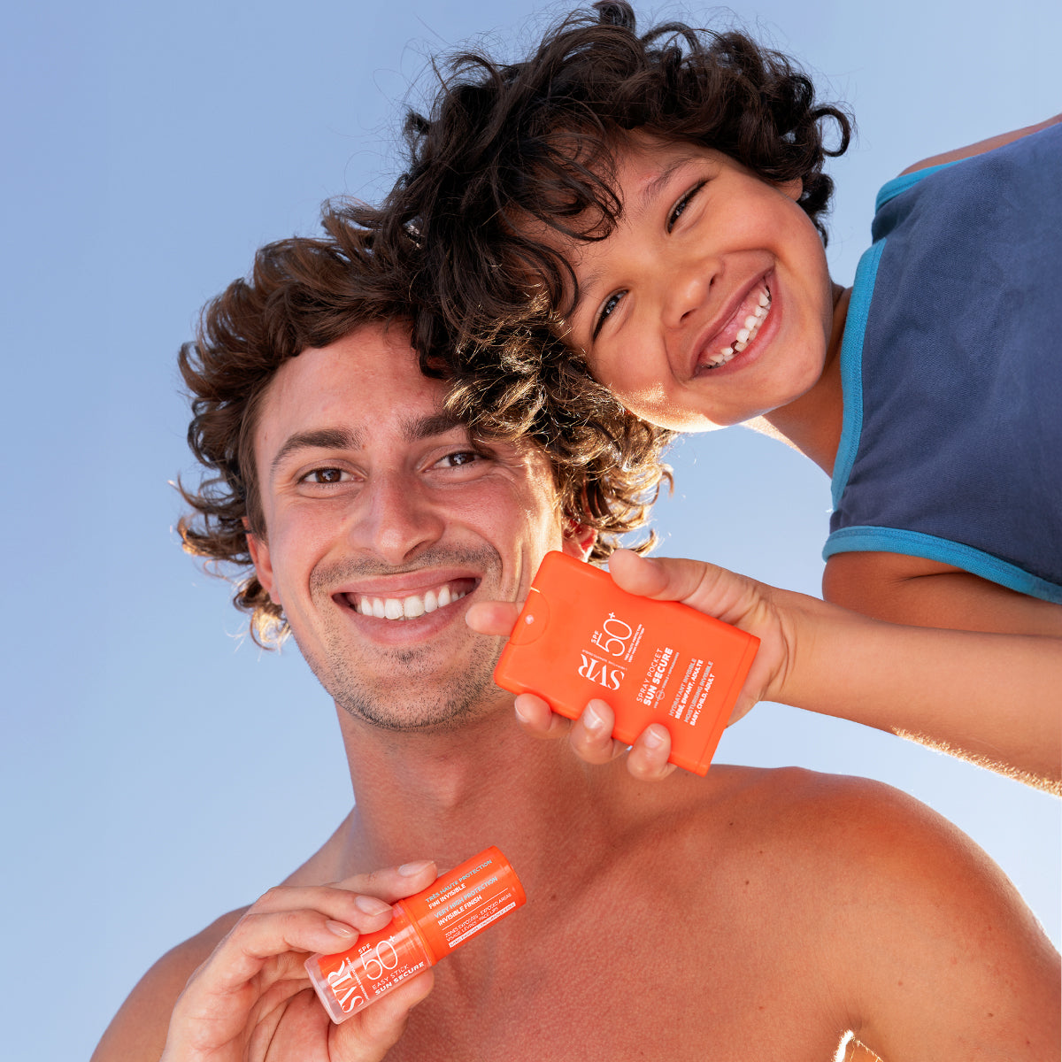 Smiling man and child holding orange SVR SUN SECURE SPF50+ spray and stick sunscreen against a clear blue sky.