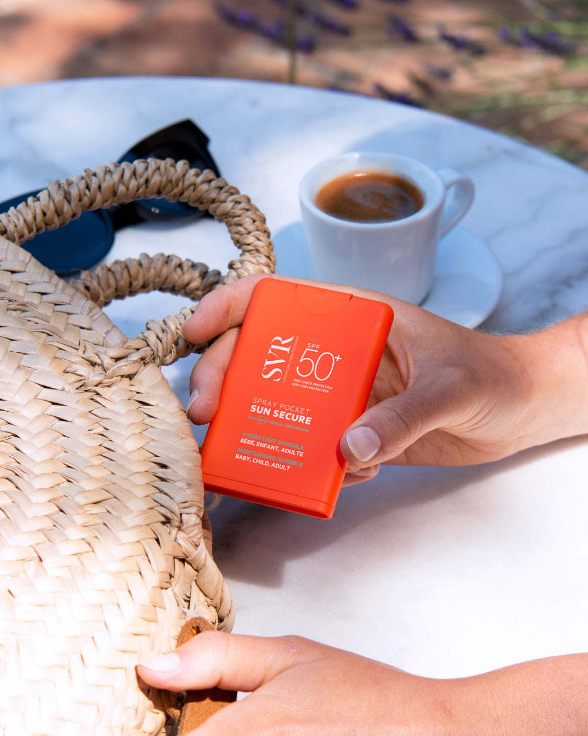 Hand holding an orange SVR SUN SECURE SPF50+ pocket spray bottle next to a straw bag and a cup of coffee on a table.