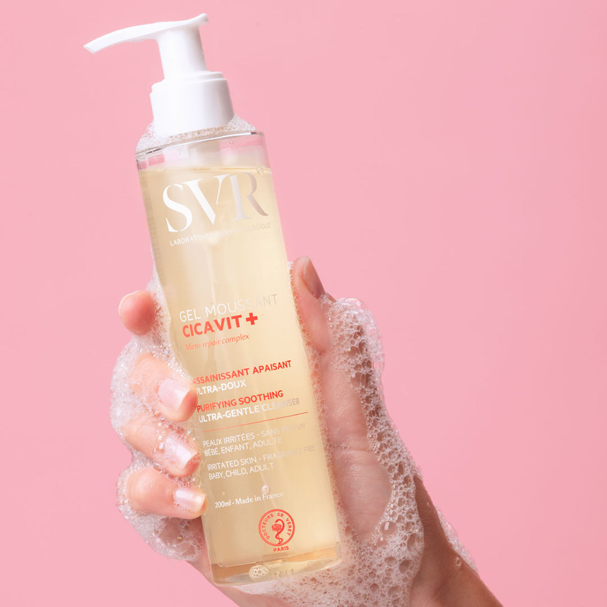 Flacon pompe de gel moussant SVR Cicavit+ tenu en main avec une mousse nettoyante sur fond rose.