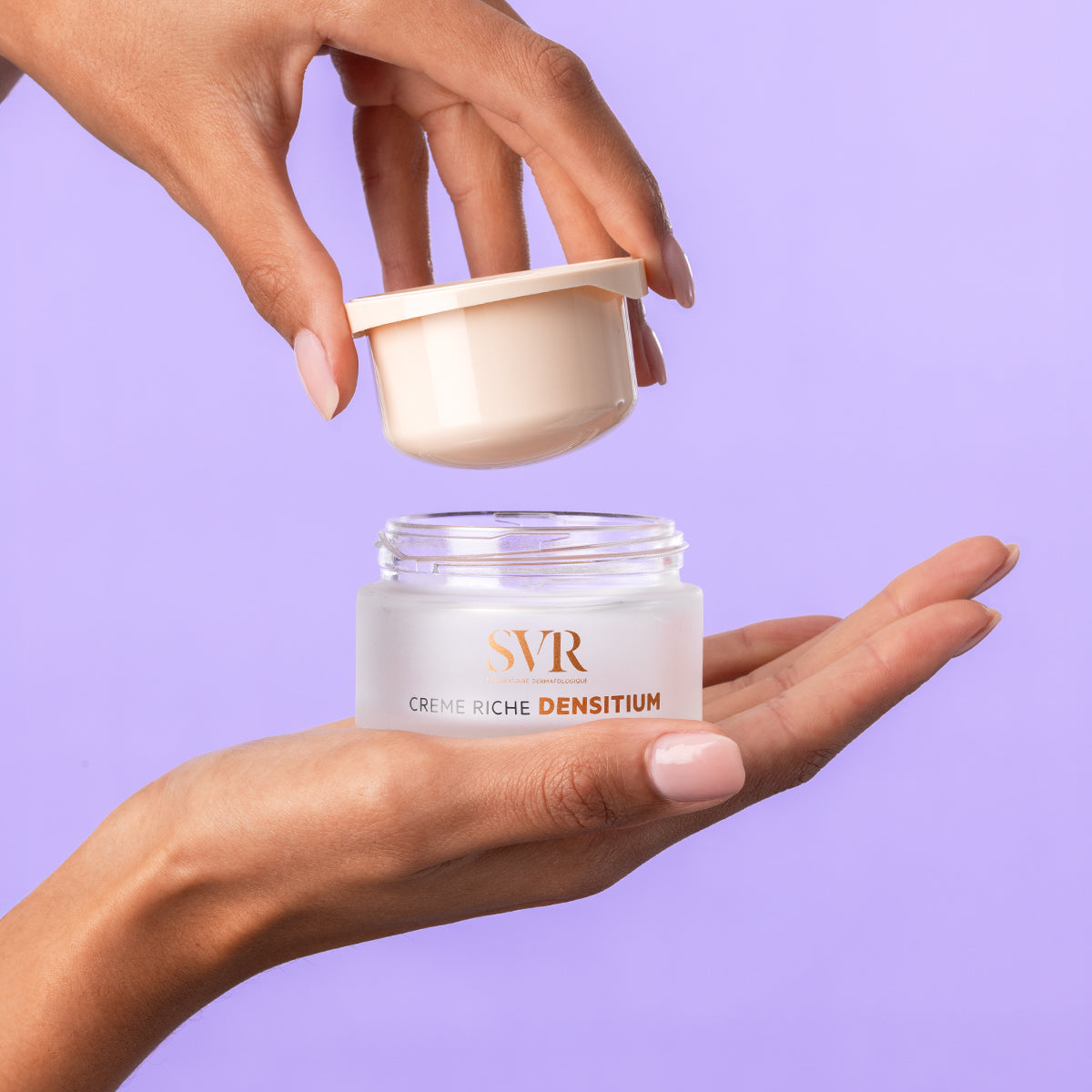 Hands holding an open jar of SVR DENSITIUM Crème riche with a beige lid against a light purple background.