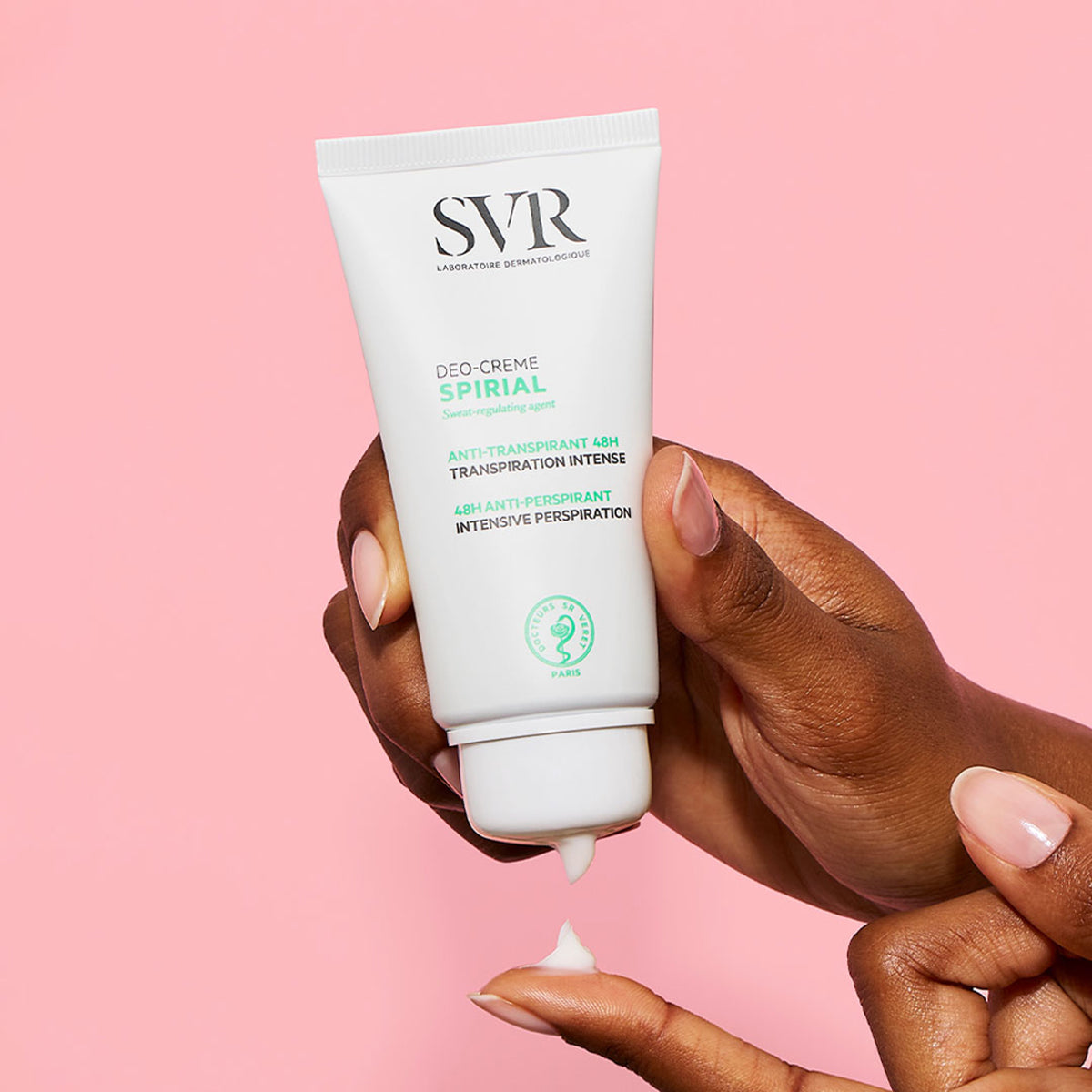 Tube de crème anti-transpirante SVR Spirial blanc tenu en main, déposant une texture crème sur le doigt, fond rose.