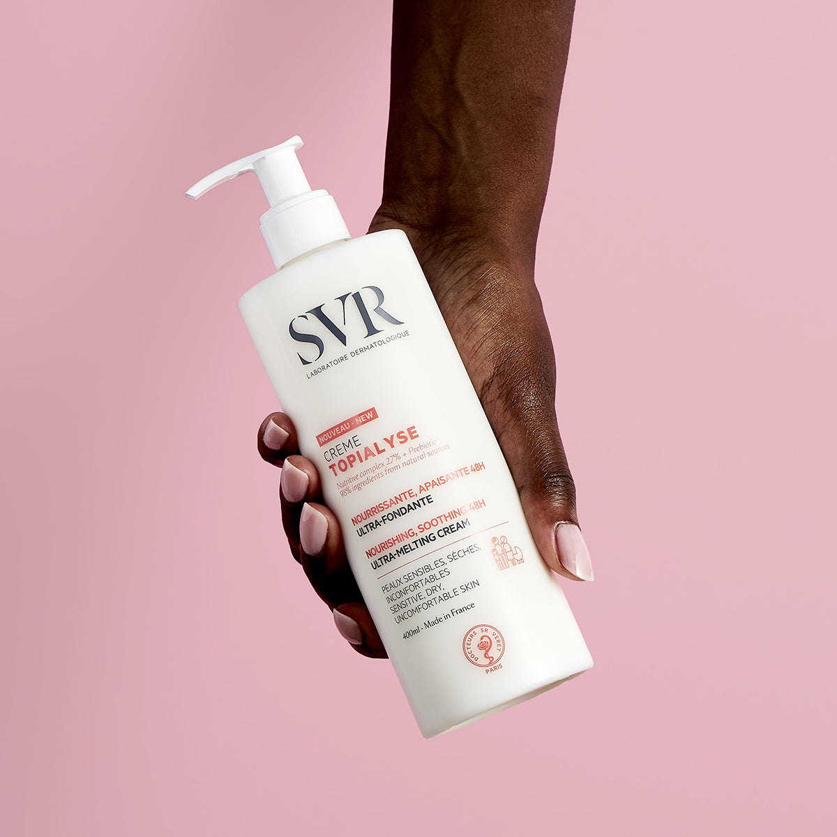 Main tenue par une main foncée présentant un flacon pompe blanc de crème hydratante SVR Topialyse sur fond rose.