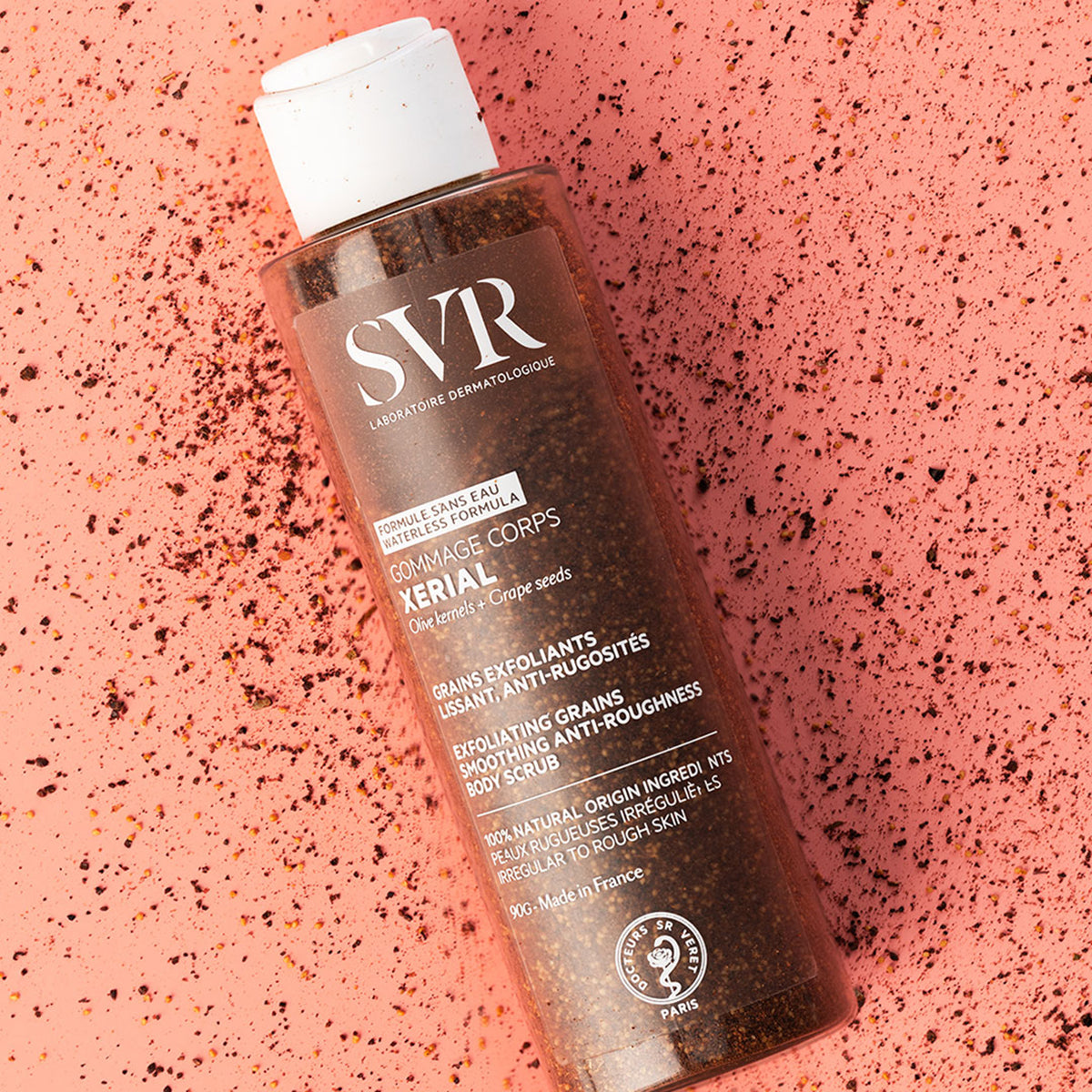 Flacon de gommage corporel SVR Xerial avec grains exfoliants naturels sur un fond texturé rose parsemé de résidus de noyaux.