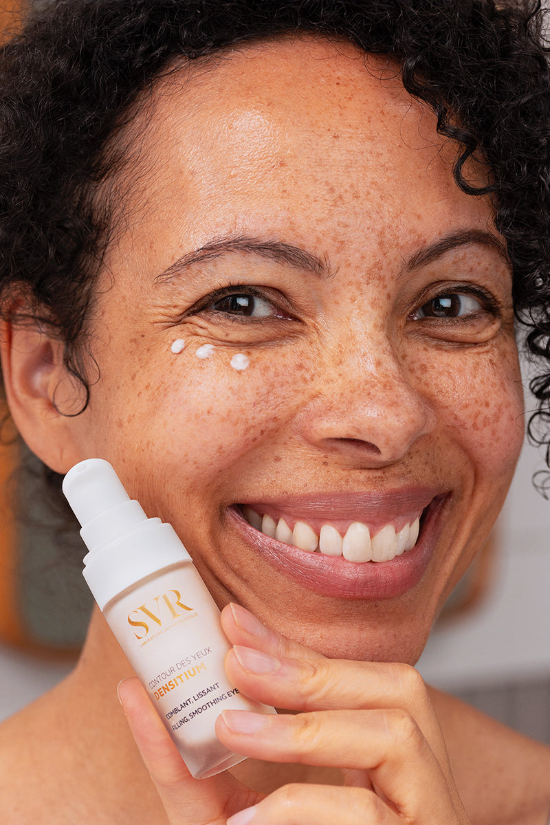 Femme souriante appliquant la crème SVR Densitium Contour des Yeux blanche avec flacon pompe en main.
