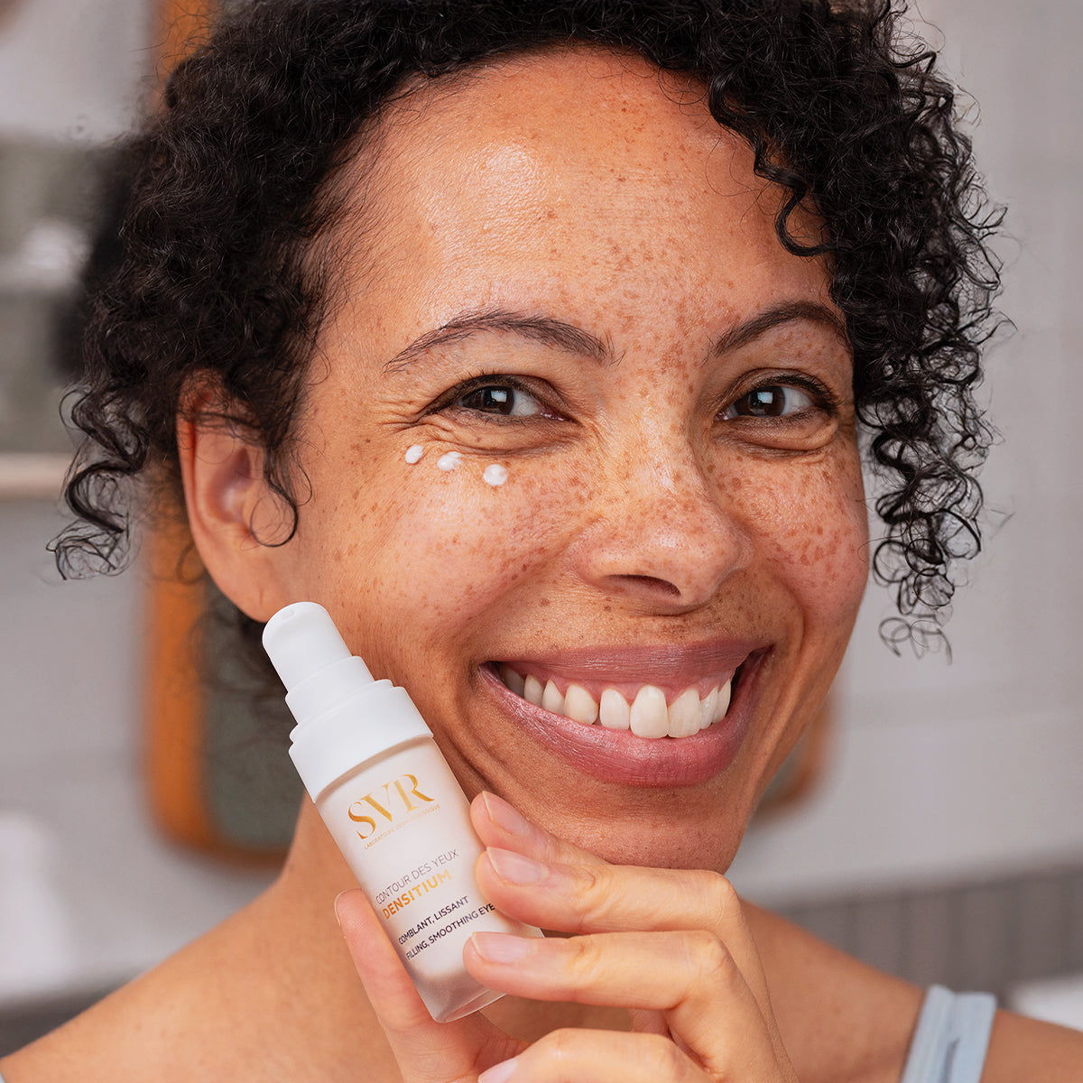 Femme souriante appliquant la crème SVR Densitium Contour des Yeux blanche avec flacon pompe en main.