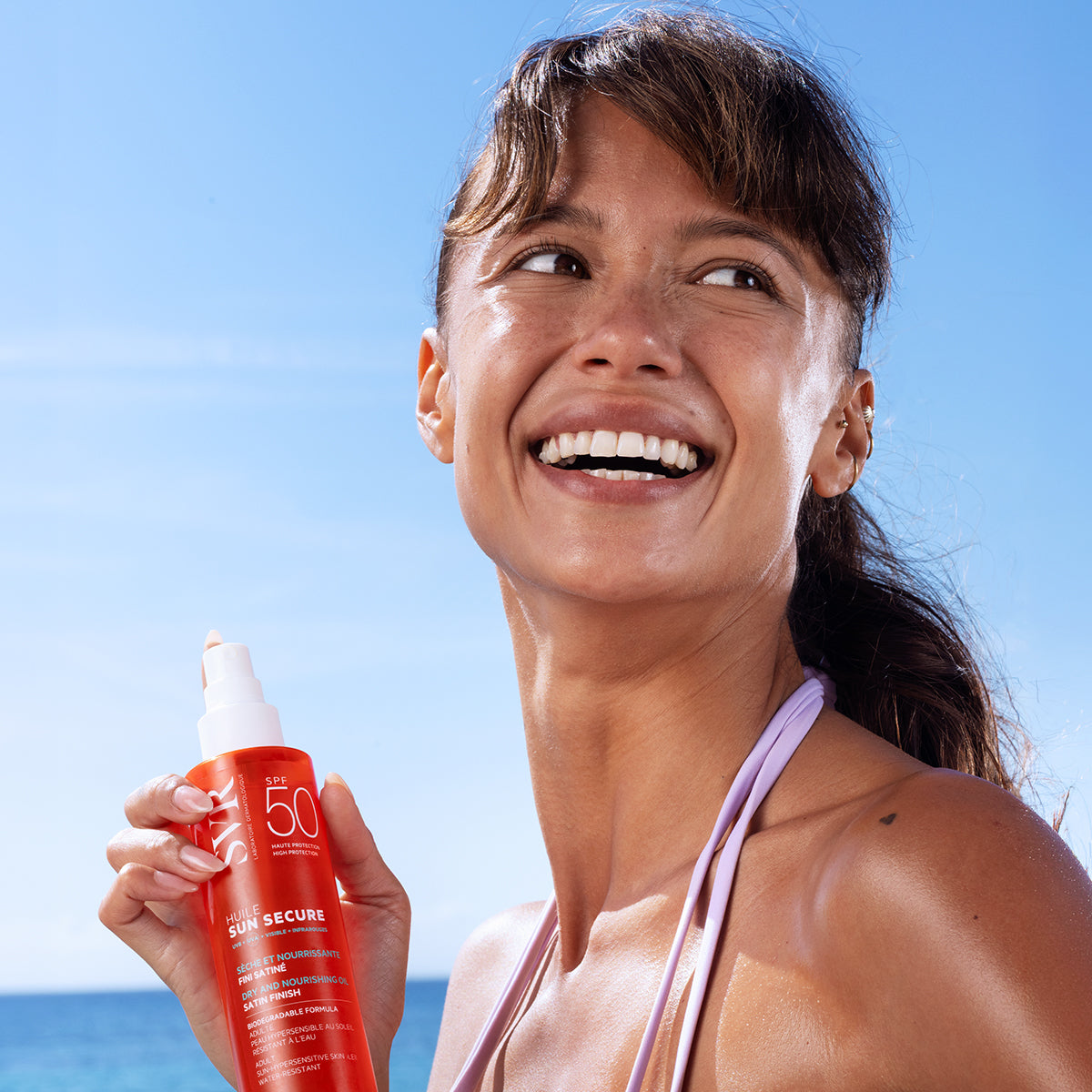 Femme souriante tenant un flacon orange de SVR SUN SECURE Huile SPF50 sur une plage ensoleillée.