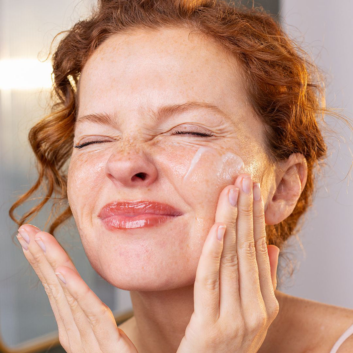 Femme rousse aux yeux fermés appliquant le baume démaquillant SVR Sensifine sur son visage pour un nettoyage doux.