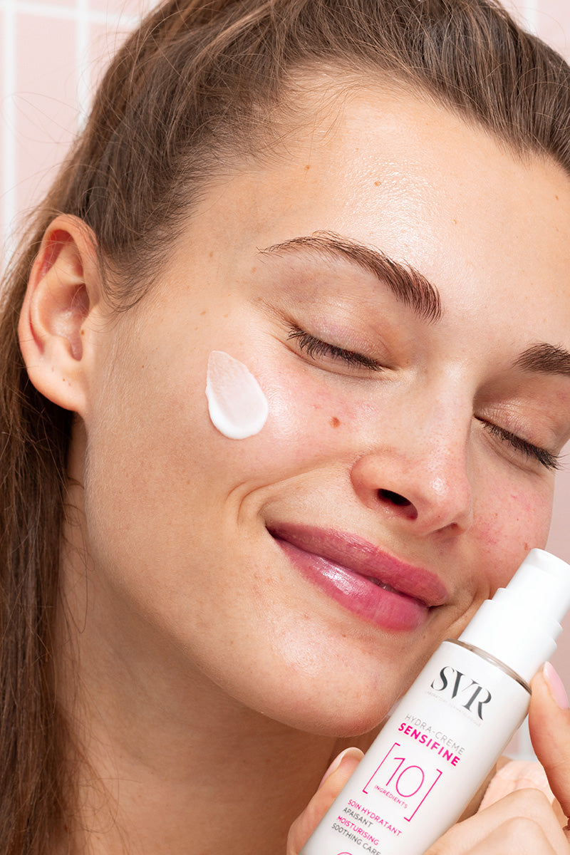 Woman applying white SVR Sensifine Hydra-Crème to her cheek in front of a pink tiled wall.