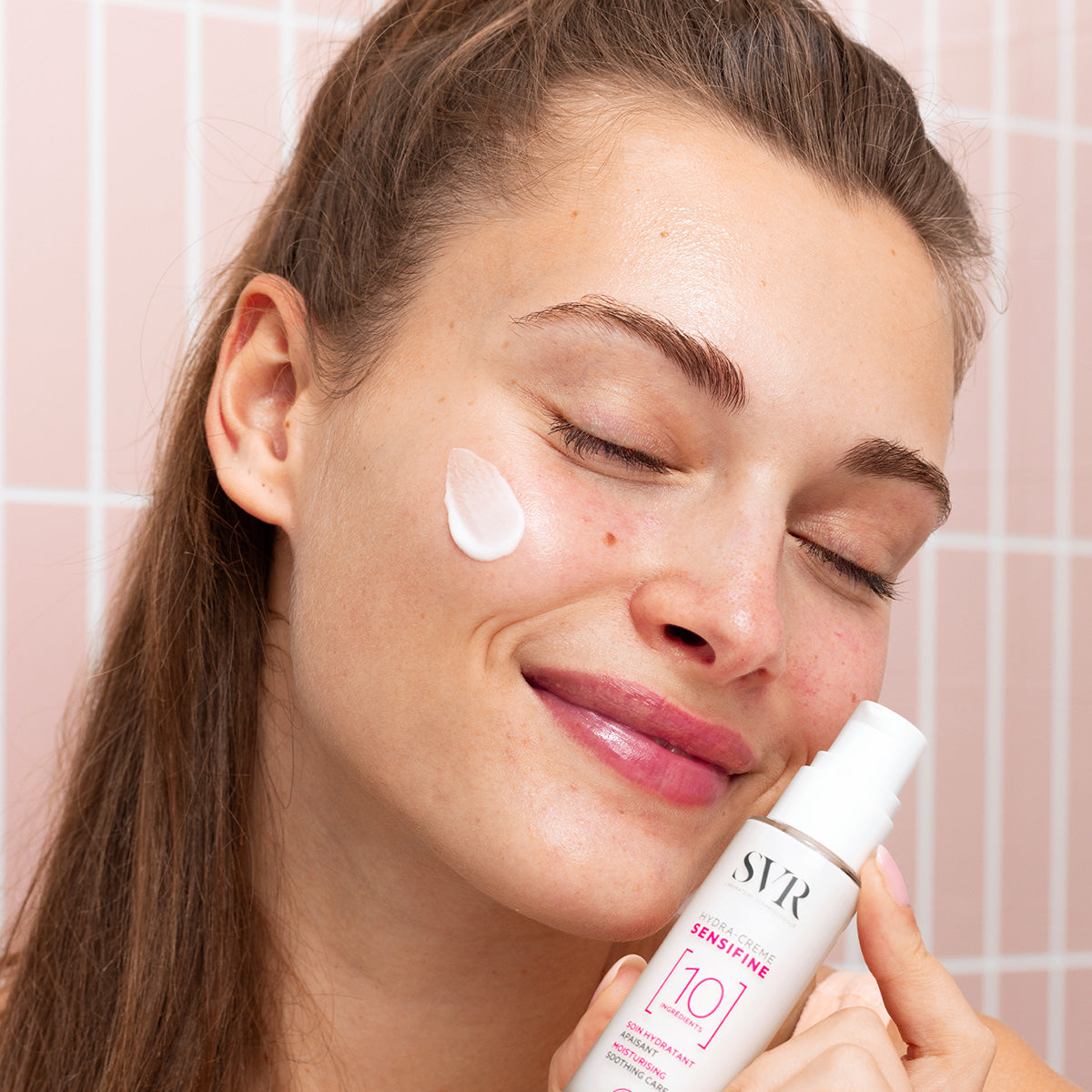 Woman applying white SVR Sensifine Hydra-Crème to her cheek in front of a pink tiled wall.