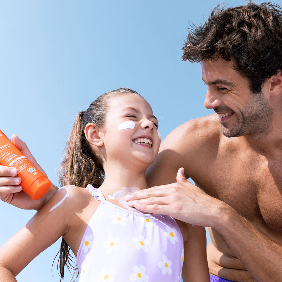 Père appliquant le lait solaire SVR Sun Secure SPF50+ sur les épaules de sa fille souriante sous un ciel bleu.