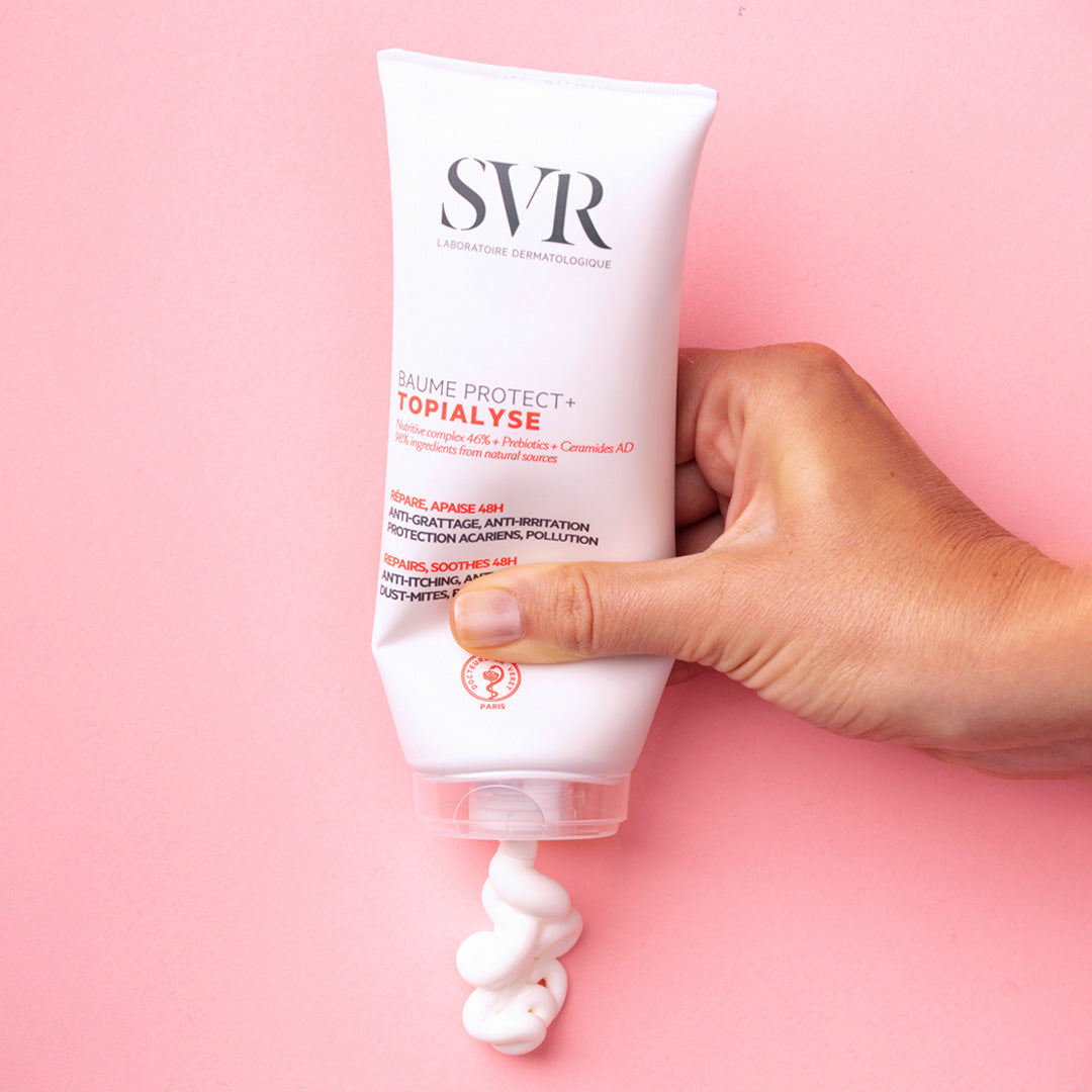 Main tenue pressant un tube blanc de baume SVR Topialyse Protect+ laissant sortir une texture crème sur fond rose.