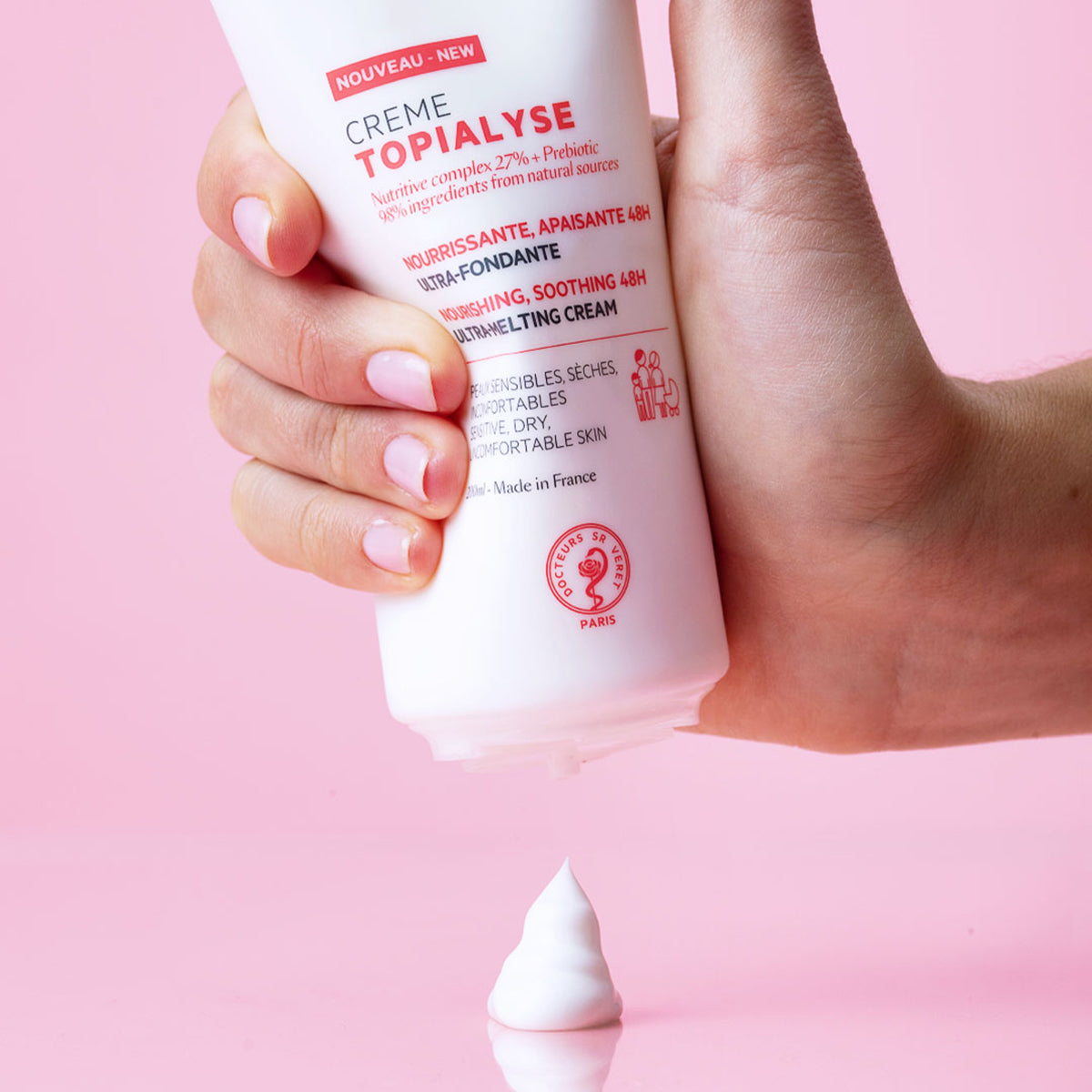 Main tenue pressant un tube blanc de crème hydratante Topialyse sur fond rose, laissant échapper une noisette de produit.