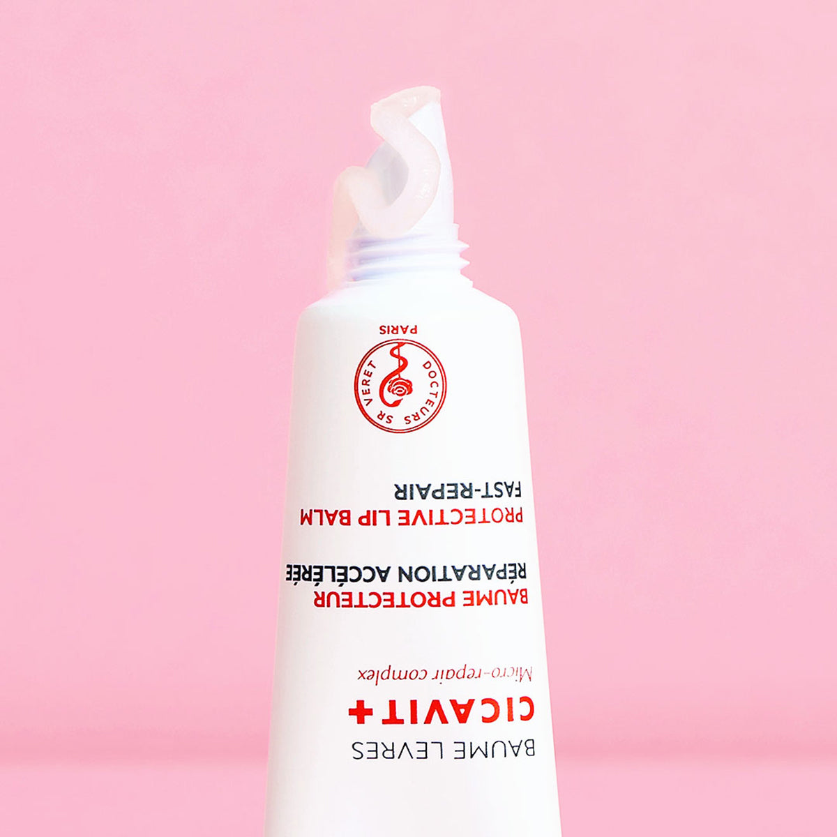 Tube blanc de baume réparateur Cicavit+ Lèvres avec une noisette de crème sur fond rose pastel.