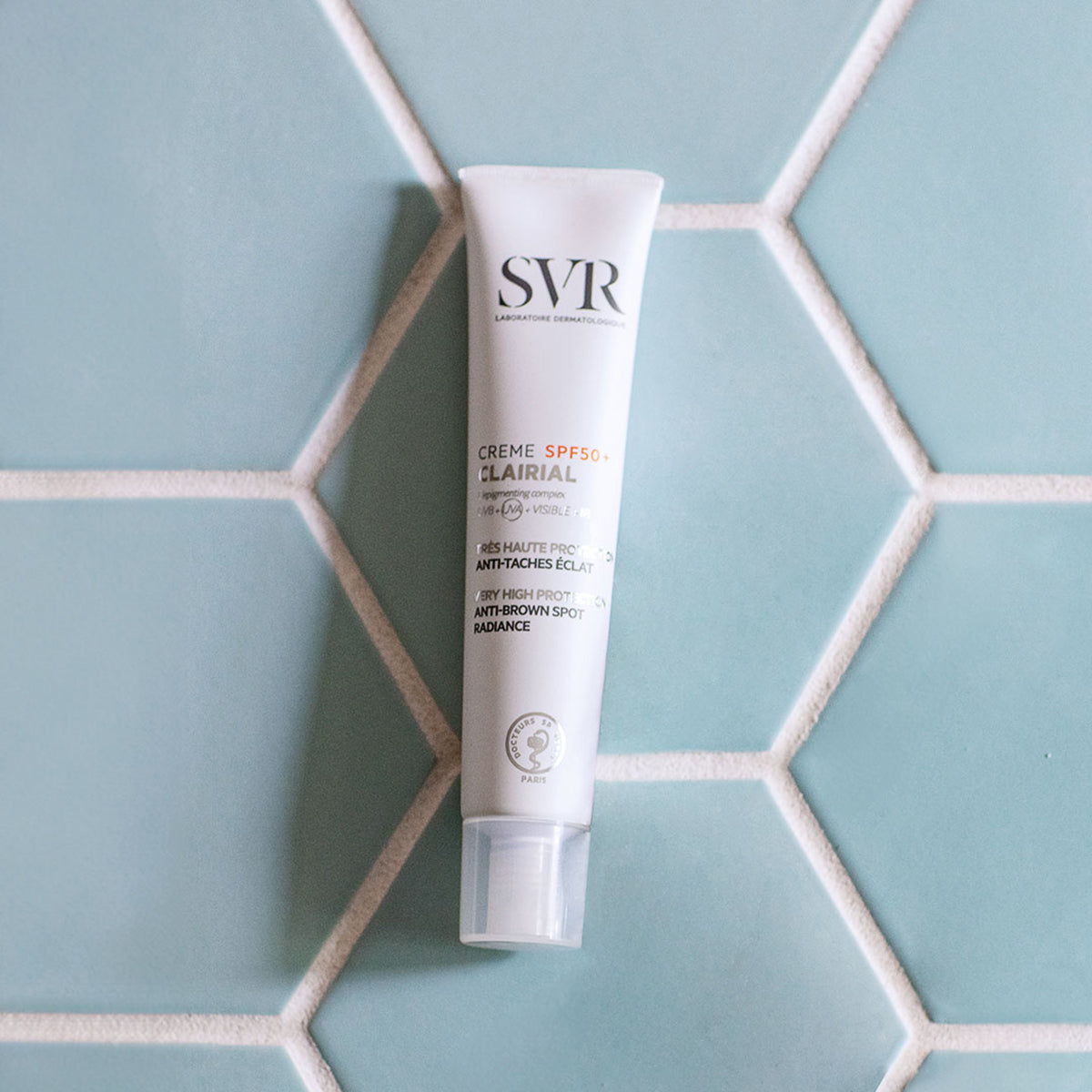 Tube blanc de crème solaire SVR Clairial SPF50+ posé sur un carrelage mural bleu clair en forme d'hexagones.