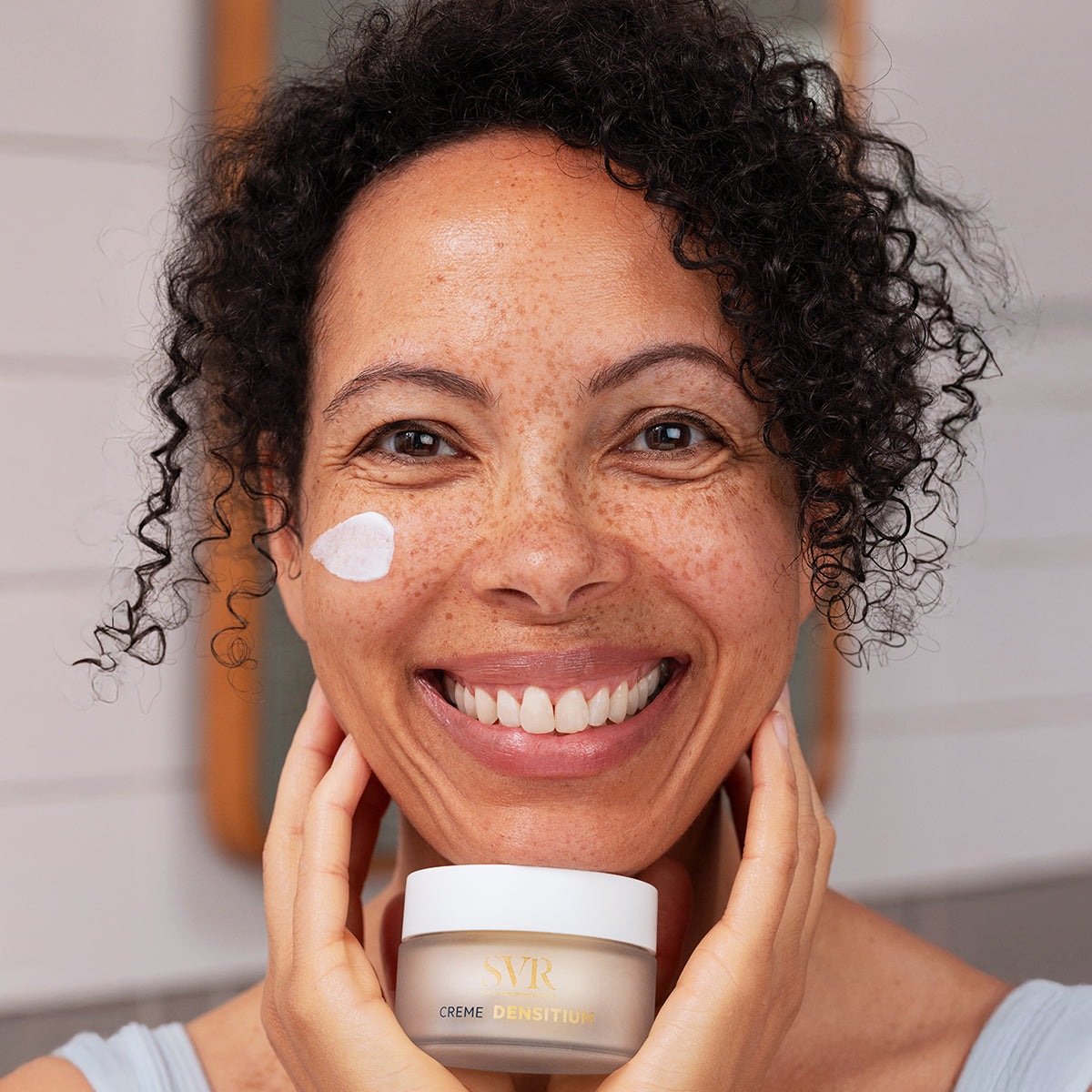 Smiling woman with curly hair holding a jar of SVR DENSITIUM Crème with a dab of white face cream on her cheek.