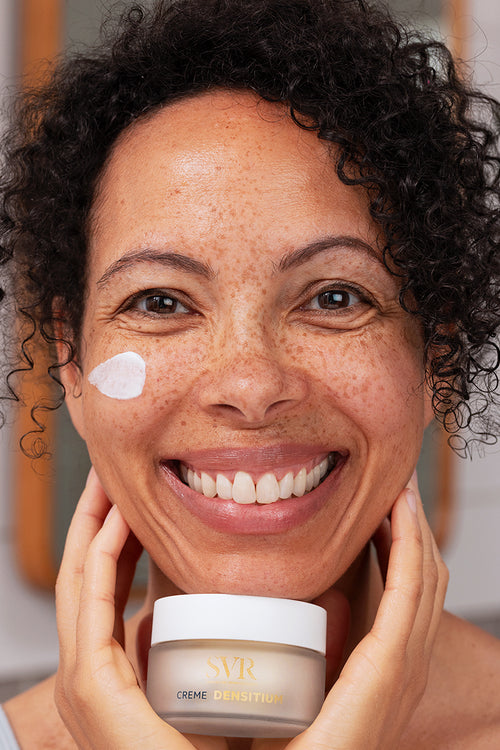 Smiling woman with curly hair holding a jar of SVR DENSITIUM Crème with a dab of white face cream on her cheek.