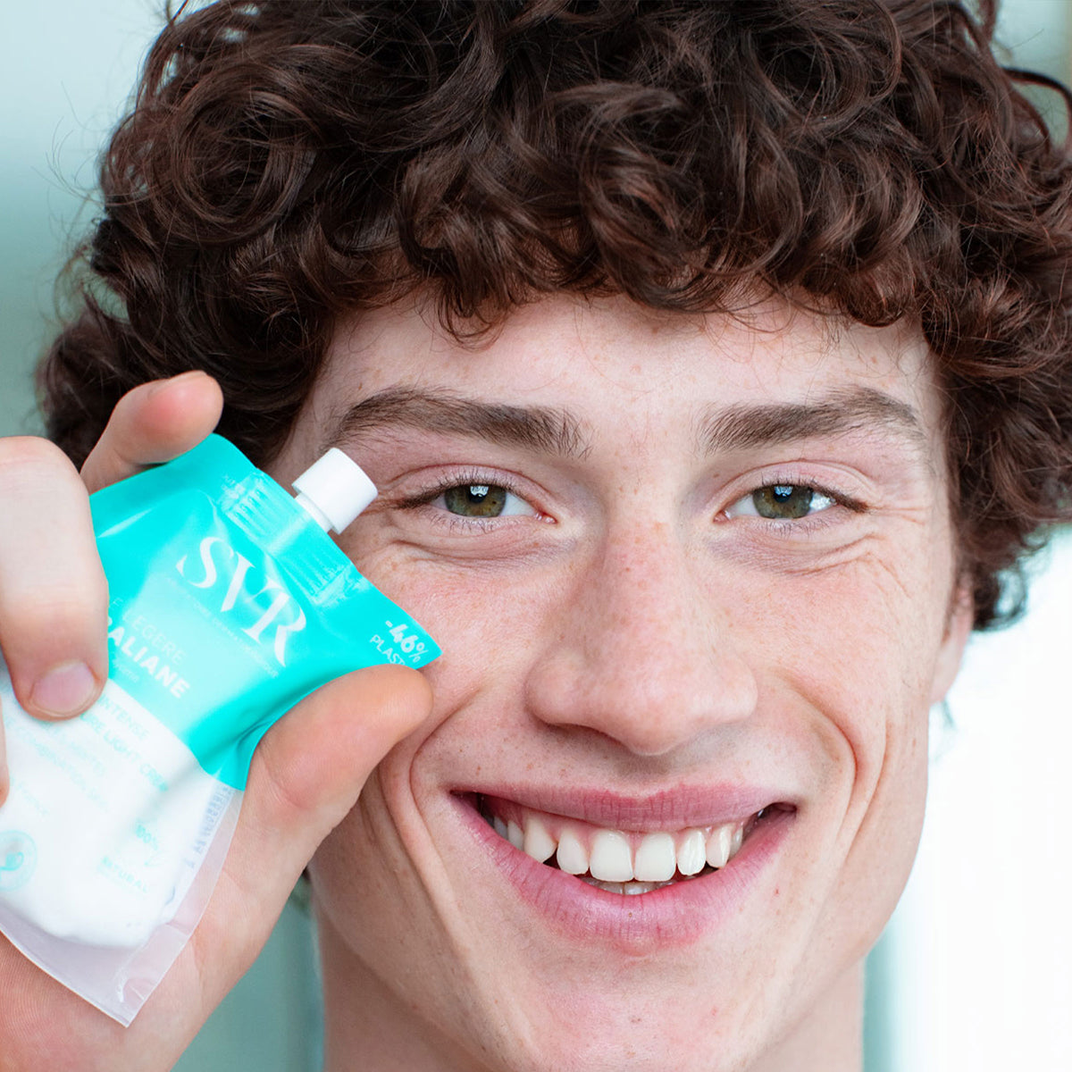 Jeune homme souriant tenant une poche éco-conçue SVR Hydraliane Crème Légère turquoise et transparente.
