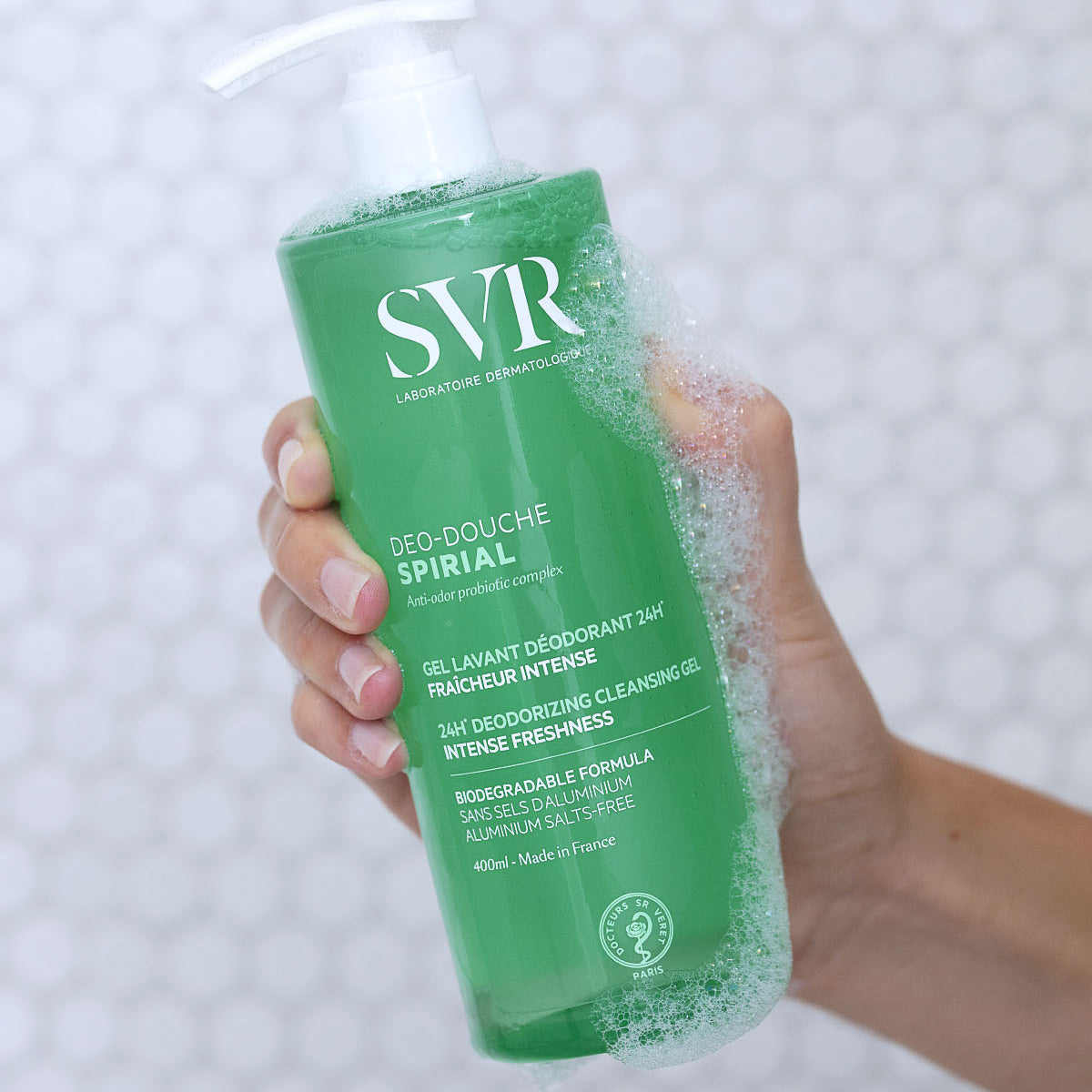 Flacon pompe vert de gel douche SVR Spirial 400ml tenu en main avec une mousse nettoyante sur le dessus.