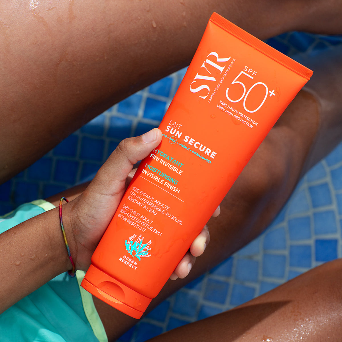 Tube de lait solaire SVR Sun Secure SPF50+ orange tenu à la main au bord d'une piscine bleue.