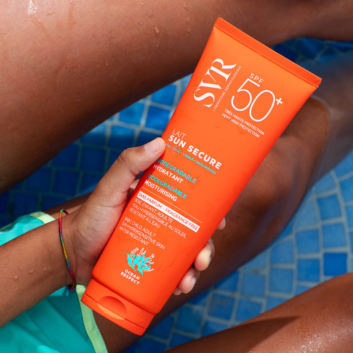 Main orange SVR SUN SECURE SPF50+ sunscreen tube held by a hand against a blue tiled pool background.