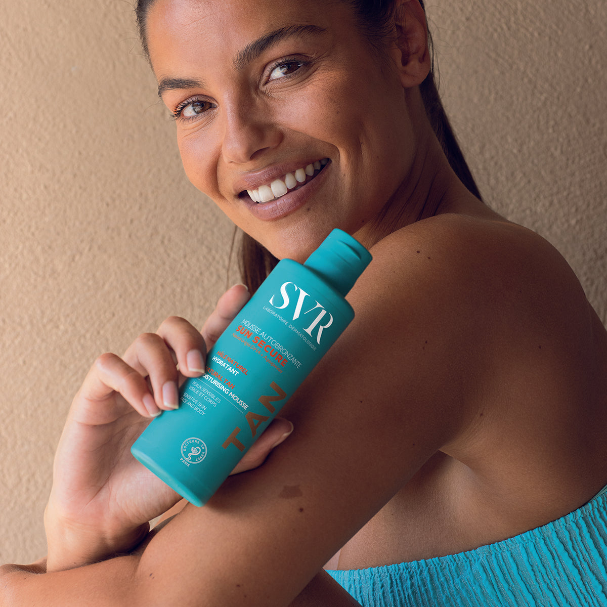 Femme souriante tenant un flacon bleu turquoise de mousse autobronzante SVR Sun Secure pour le visage et le corps.