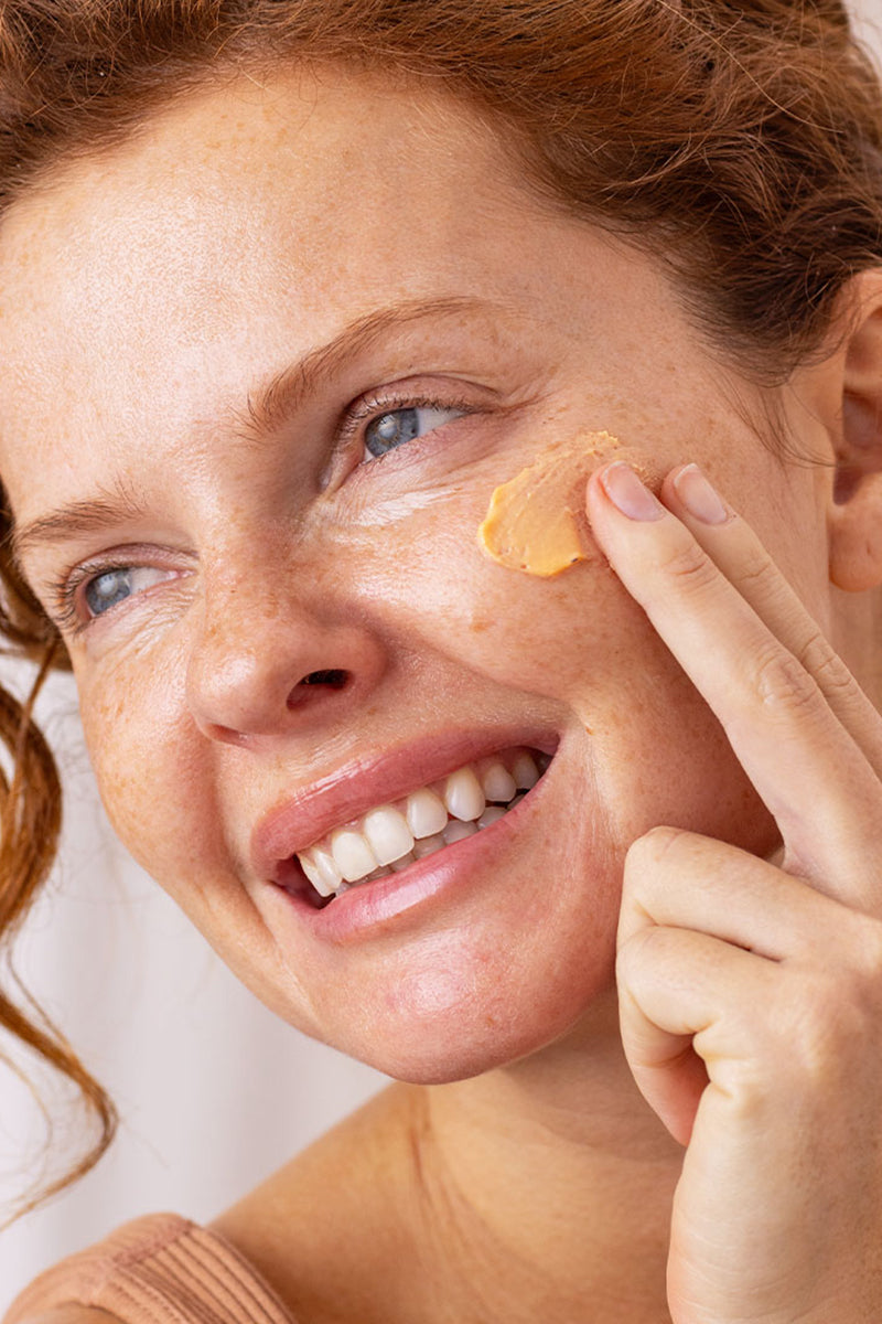 Femme rousse souriante appliquant une crème solaire mousse abricotée sur sa joue pour un effet floutant et unifié.