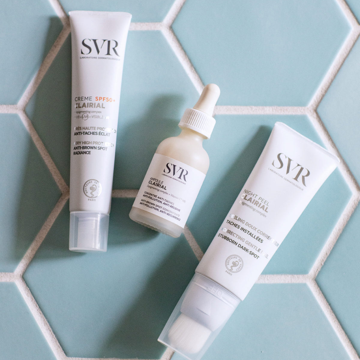 Trois produits SVR Clairial, dont une ampoule à pipette et deux tubes blancs, posés sur un carrelage bleu clair hexagonal.
