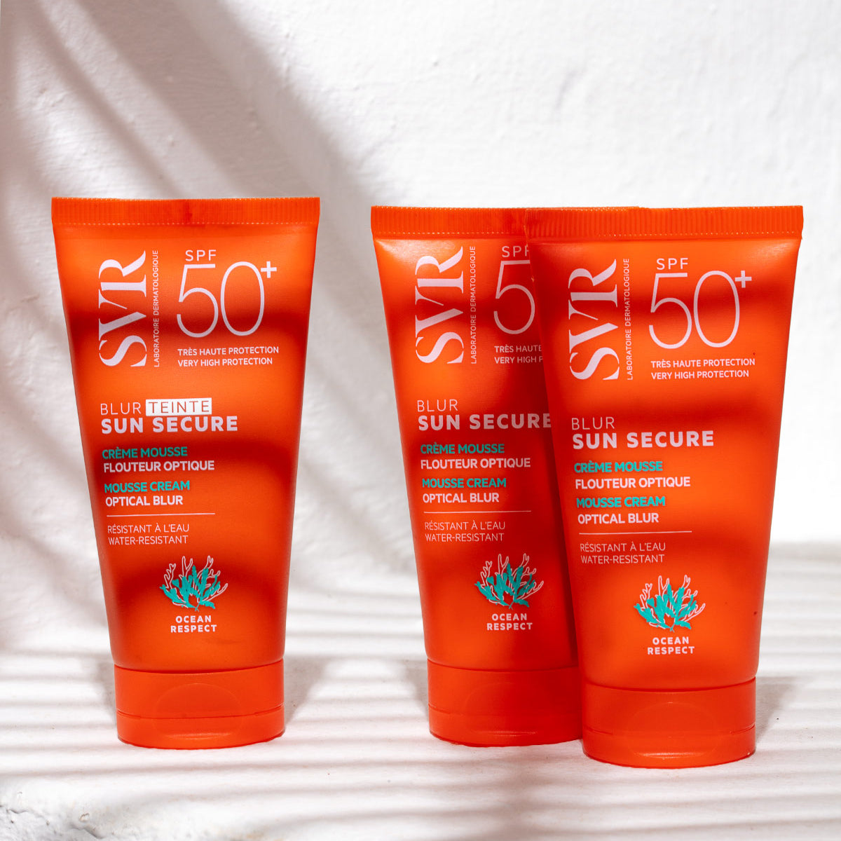 Trois tubes de crème solaire SVR Sun Secure Blur SPF50+ orange vif sur un fond blanc texturé avec des ombres de feuilles.