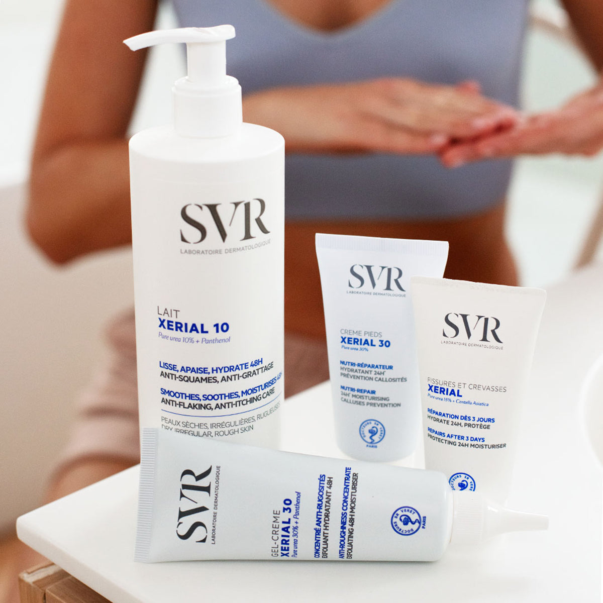Gamme de soins SVR Xerial : flacon pompe blanc et tubes de crèmes hydratantes sur une table, avec une personne en arrière-plan.
