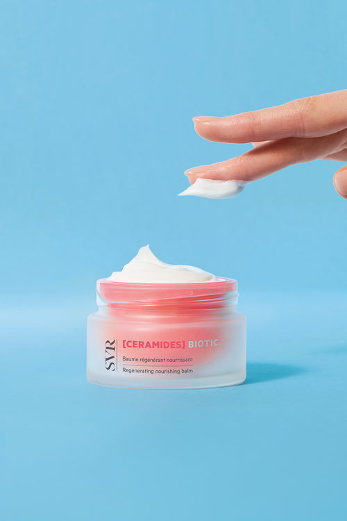 Finger applying white SVR Ceramides Biotic regenerating nourishing balm from a frosted glass jar on a blue background.
