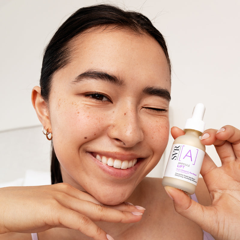 Smiling woman holding a glass bottle of SVR Ampoule Lift retinol serum with a white dropper cap.