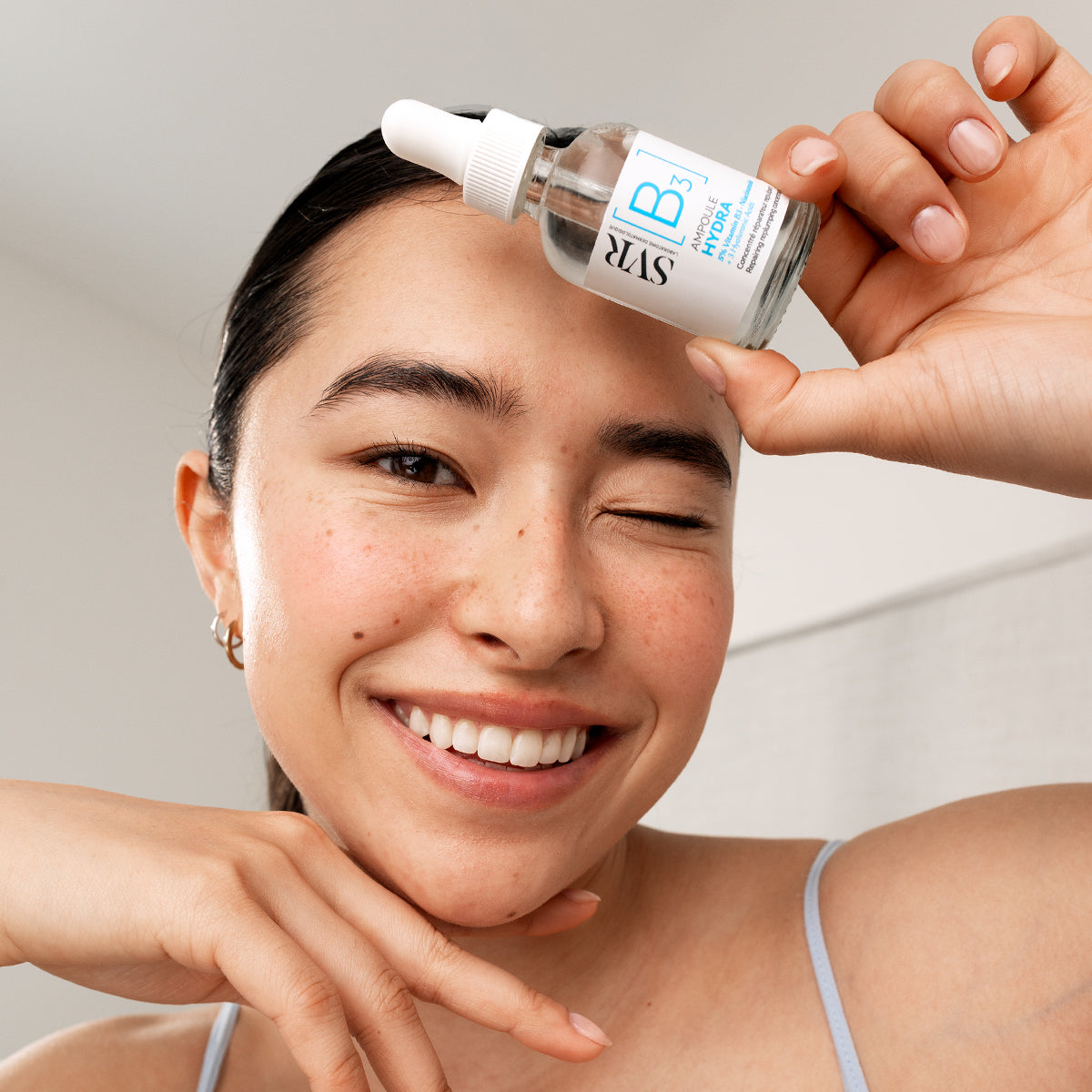 Smiling woman holding a clear glass SVR Ampoule Hydra B3 serum bottle with a white dropper cap against her forehead.