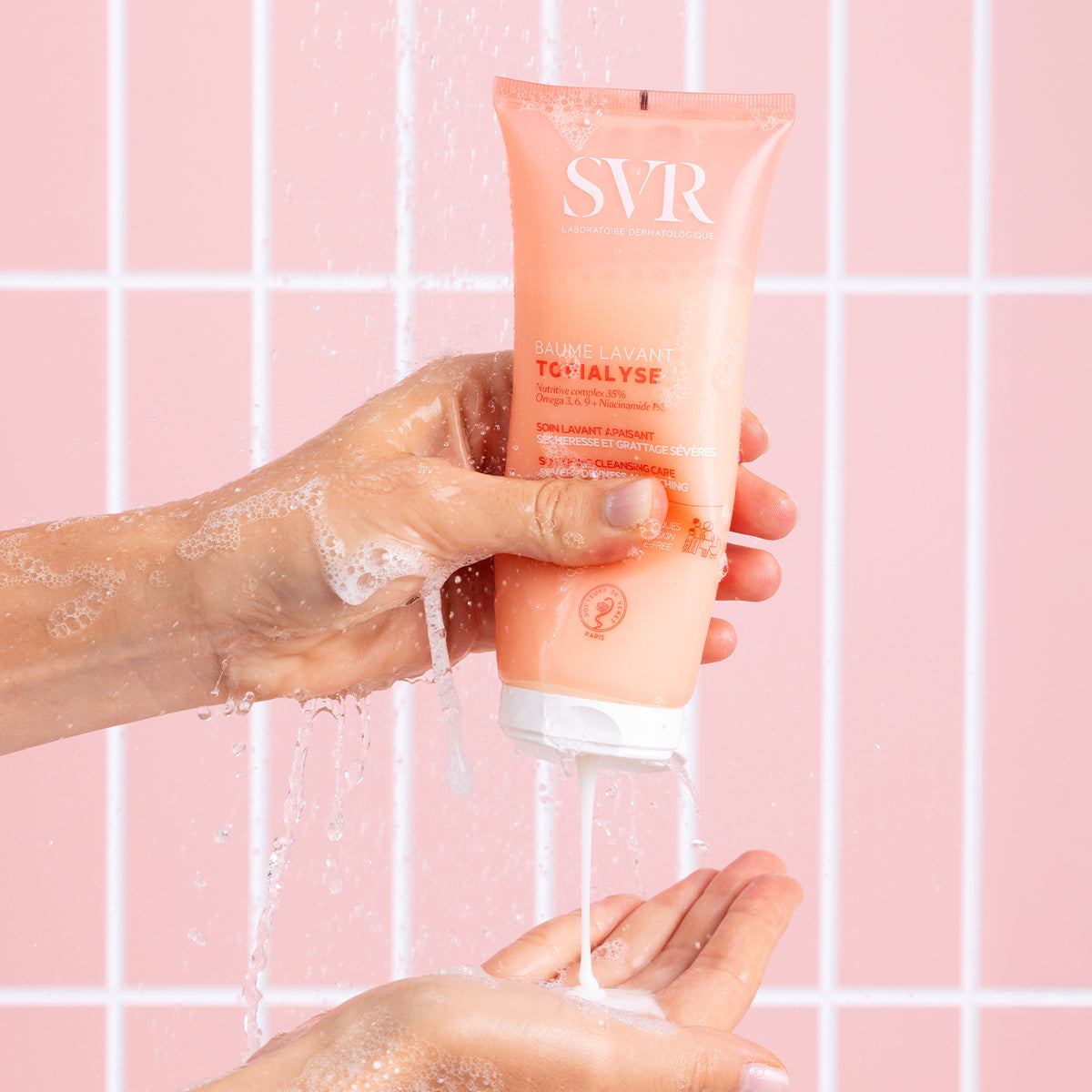 Tube de SVR Topialyse Baume Lavant rose pressé par une main sous l'eau, sur fond de carrelage rose.